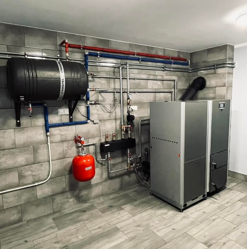 Nowoczesna kotłownia z piecem na paliwo stałe, zbiornikiem akumulacyjnym Kospel i instalacją CO w rurach miedzianych, na tle szarej, ceramicznej okładziny ściennej.