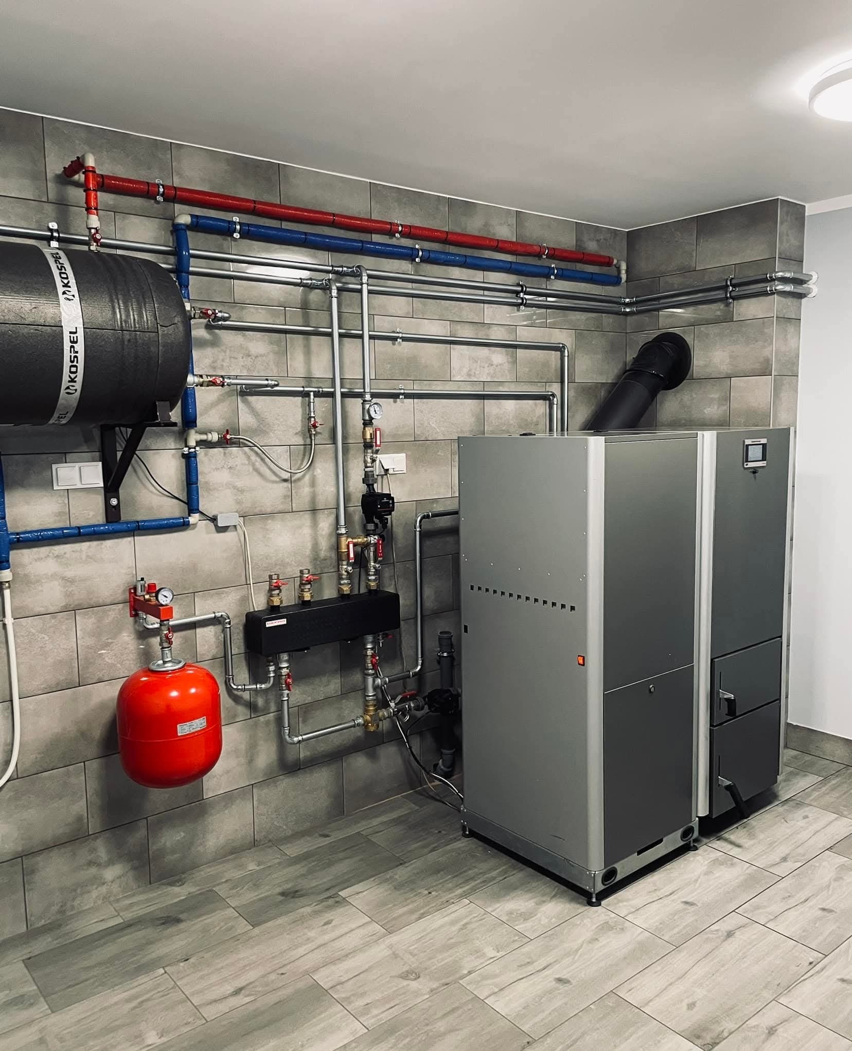 Nowoczesna kotłownia z szarymi płytkami, piecem CO, zbiornikiem buforowym Kospel i instalacją grzewczą z rurami w kolorach czerwonym i niebieskim.