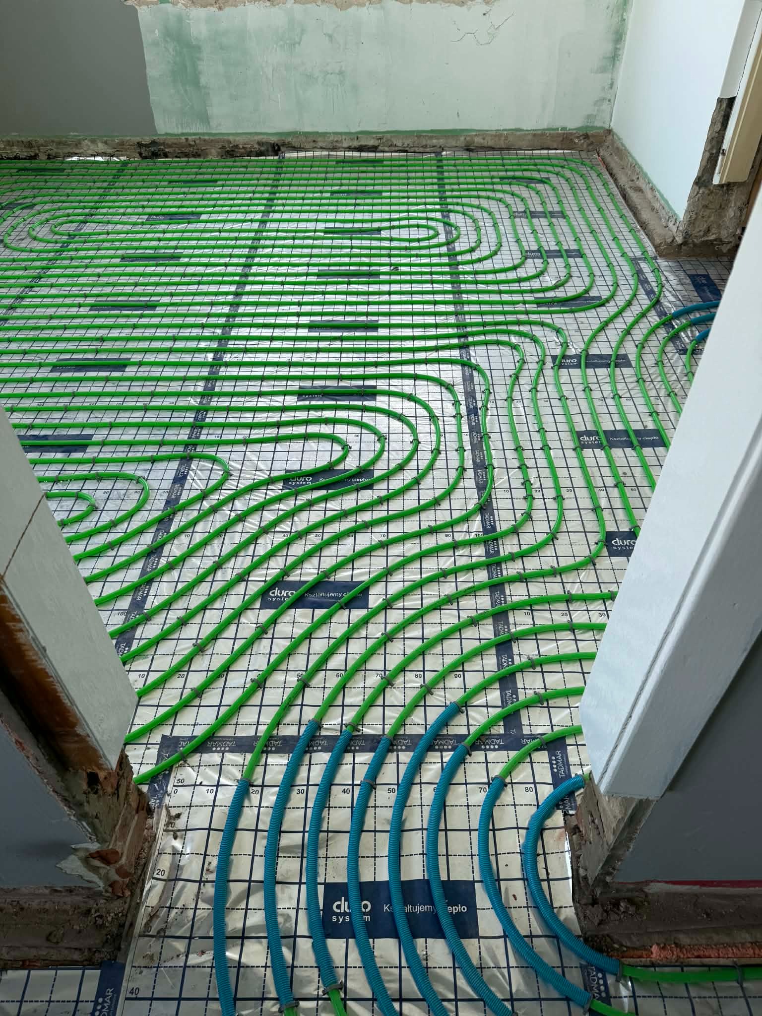 Zielone i niebieskie rury ogrzewania podłogowego ułożone na folii z nadrukiem, widoczne surowe ściany i fragment futryny drzwiowej.