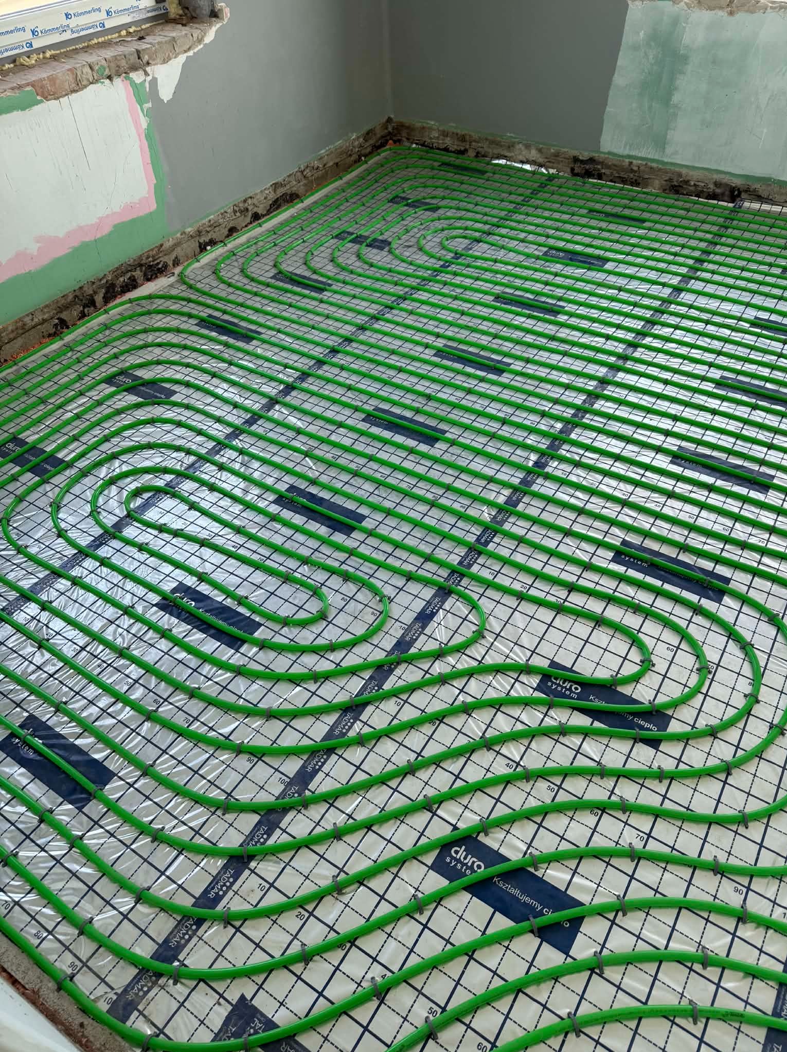 Zielone rury ogrzewania podłogowego ułożone na folii z nadrukiem siatki i logo 'duro system' w surowym wnętrzu przed wylewką. Widok z góry.