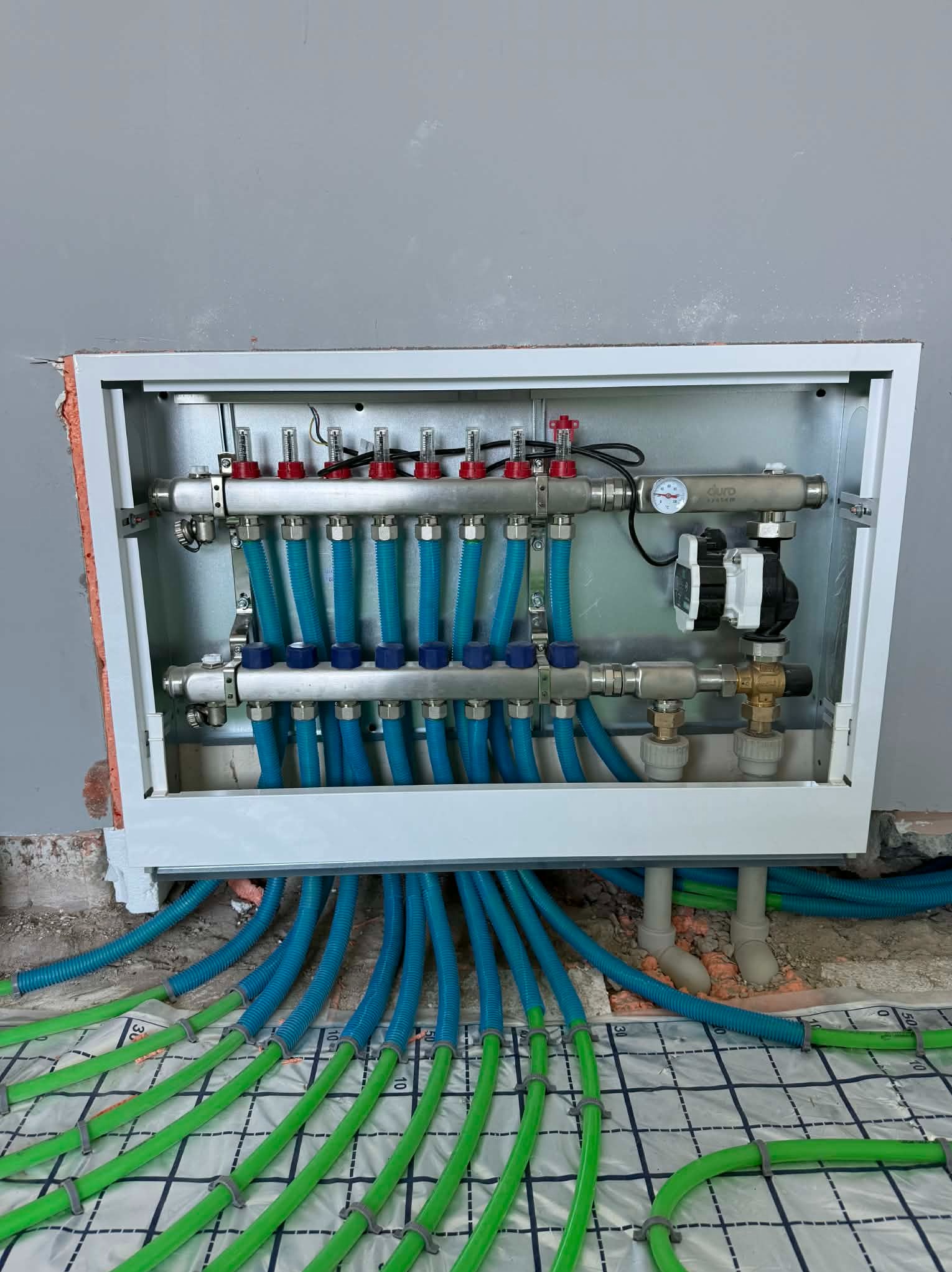 Rozdzielacz ogrzewania podłogowego z niebieskimi rurami w metalowej szafce, widoczne zielone rury ogrzewania podłogowego na podłodze z folią.