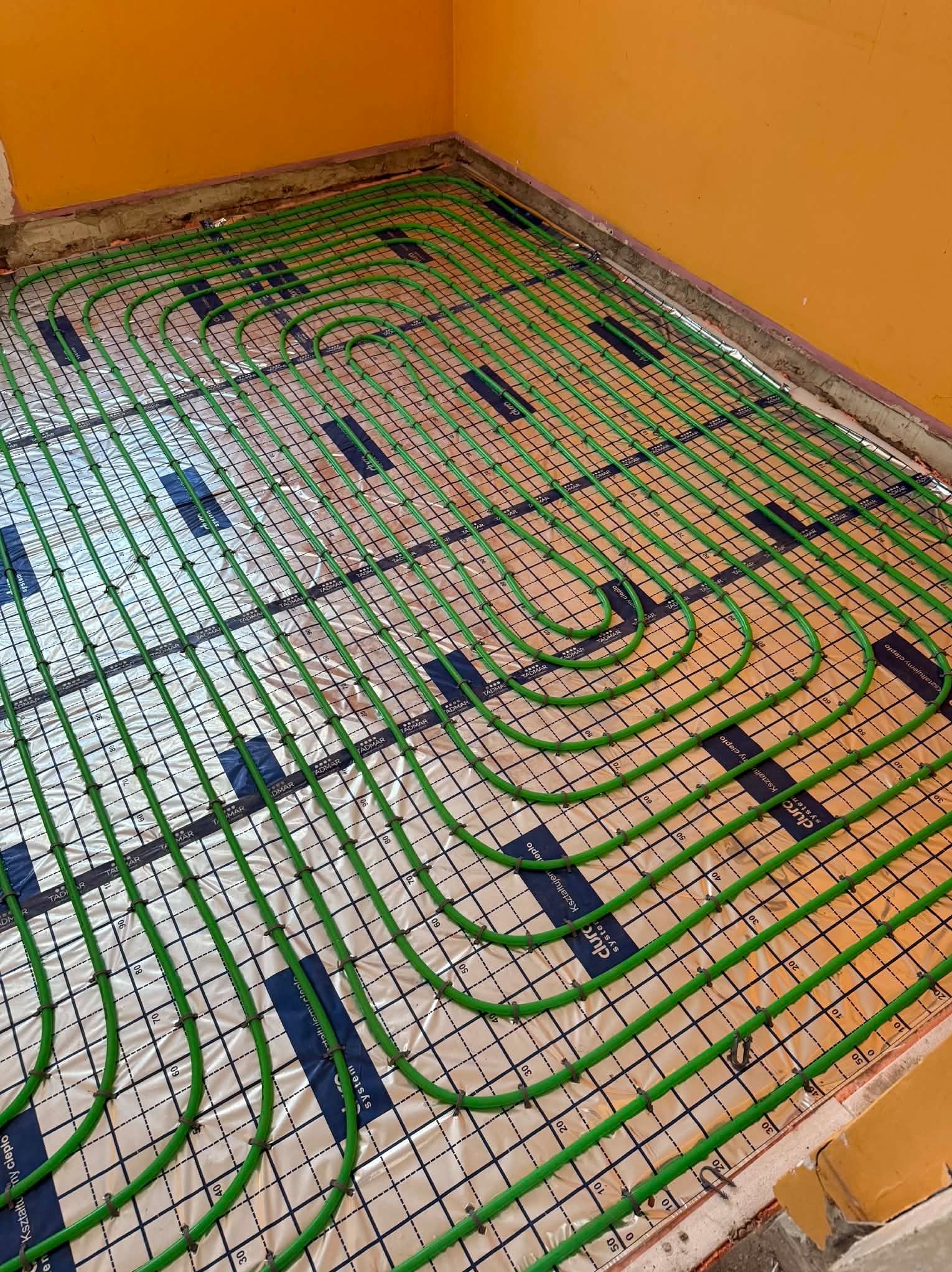Ogrzewanie podłogowe z zielonych rur na folii aluminiowej, ułożone w pętle na podłodze w Kazimierzy Wielkiej. Widok z góry, żółte ściany w tle.