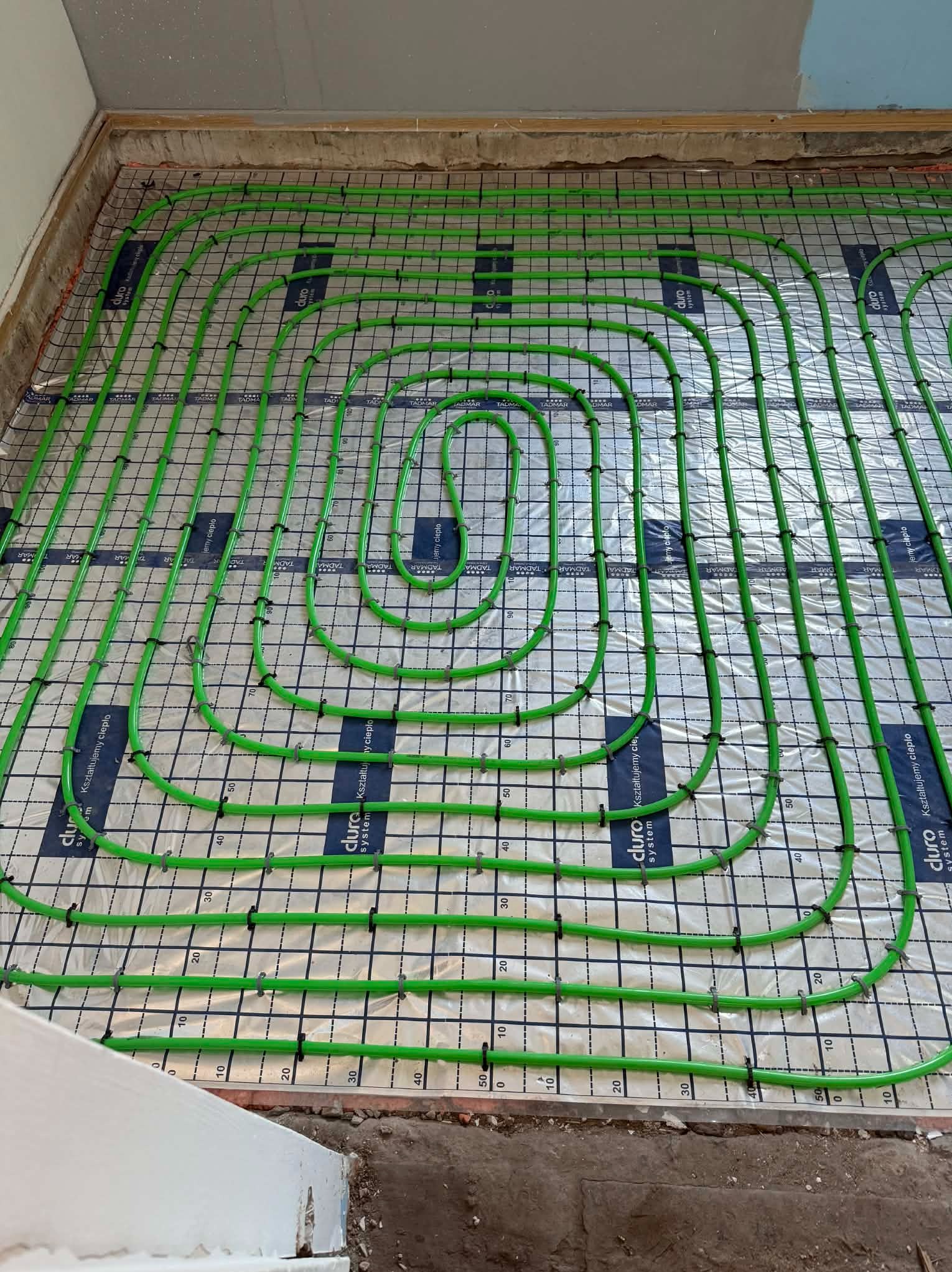 Zielone rury ogrzewania podłogowego ułożone spiralnie na folii z nadrukiem miarki, gotowe do zalania posadzką. Widok z góry na instalację w trakcie realizacji.