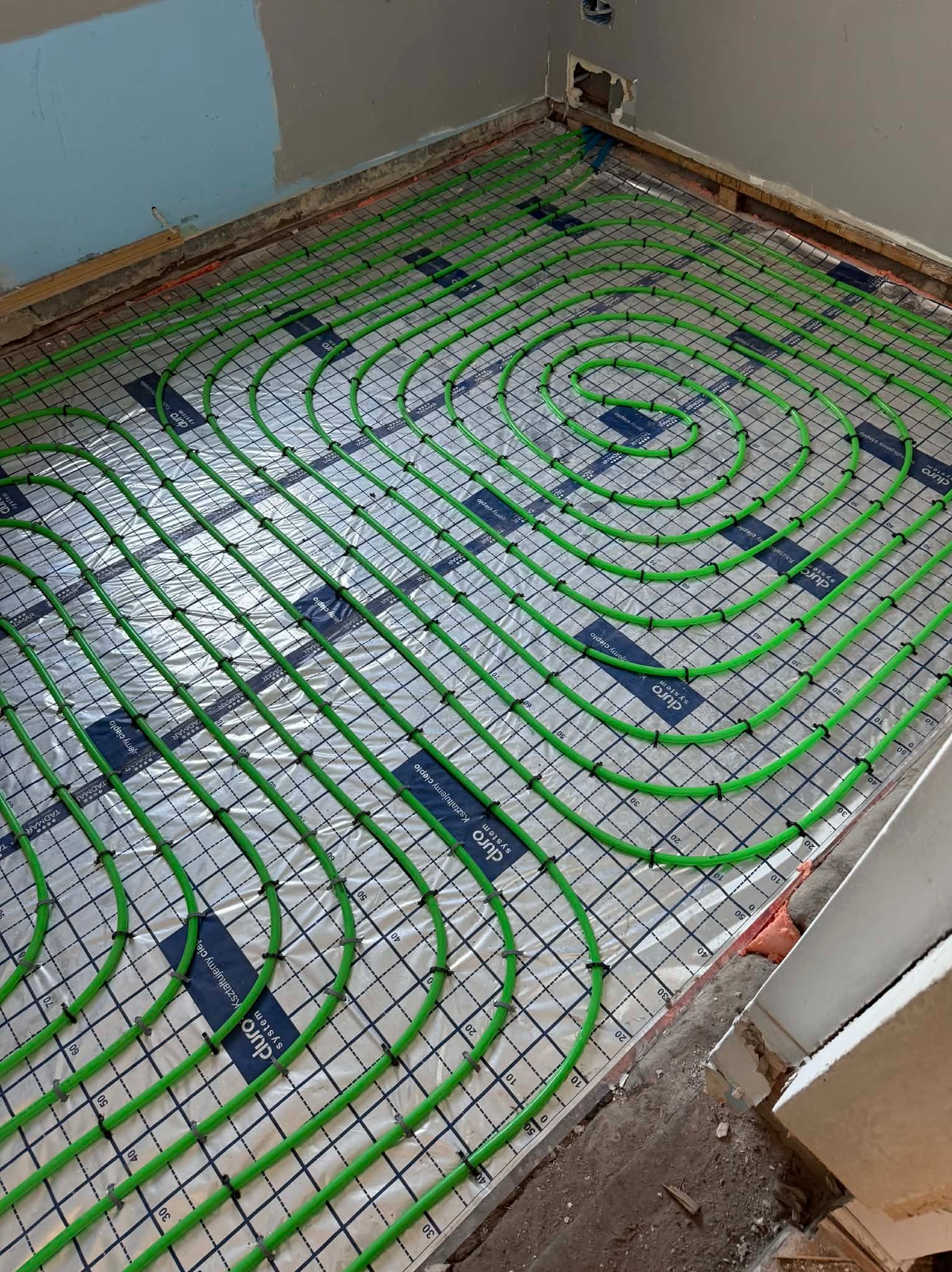 Zielone rury ogrzewania podłogowego ułożone spiralnie na folii z nadrukiem, przymocowane czarnymi uchwytami do siatki. Widok z góry na surową podłogę w trakcie instalacji.
