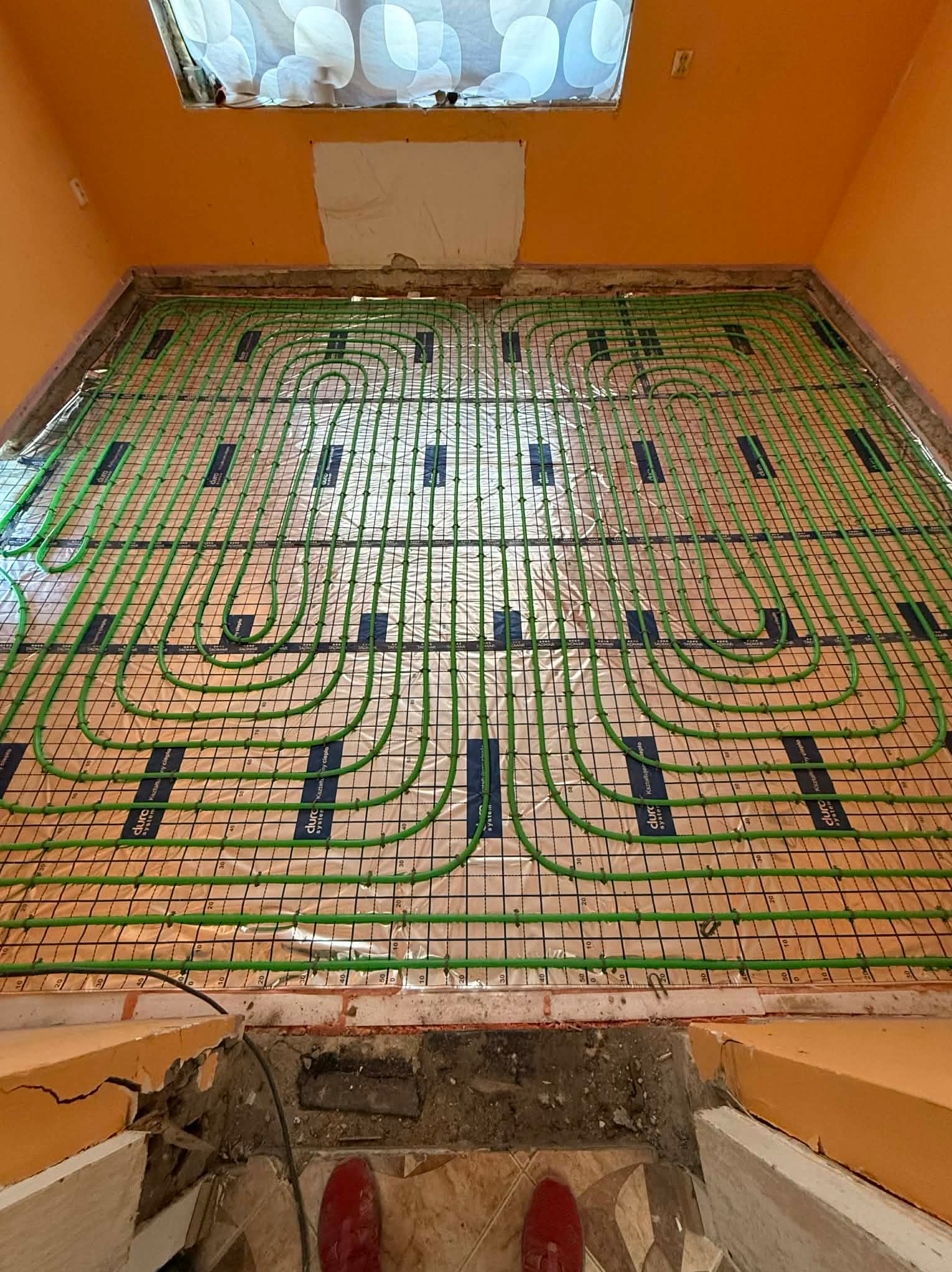 Zielone rury ogrzewania podłogowego ułożone na folii z siatką w pomarańczowym pomieszczeniu, widok z góry na instalację przed wylaniem posadzki. Widoczne czerwone buty.