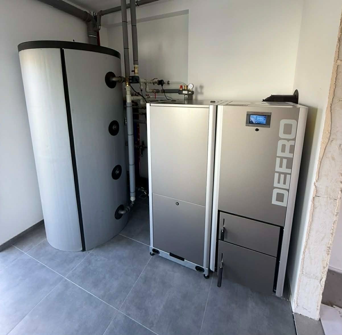 Nowoczesny, srebrny piec CO marki DEFRO z zasobnikiem na wodę, zainstalowany w kotłowni z szarymi płytkami. Widoczne rury instalacji grzewczej.