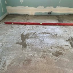 Volodymyr Trufen - Wyrównywanie podłoża w pomieszczeniu z częściowo wykończonymi ścianami. Czerwona poziomica leży na szarej, mokrej wylewce. Widoczne ślady po narzędziach.