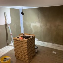 Volodymyr Trufen - Wnętrze w trakcie wykańczania: ułożone płytki podłogowe imitujące drewno, sterta kartonów z płytkami ściennymi, poziomica i narzędzia. Ściany częściowo wyszpachlowane.