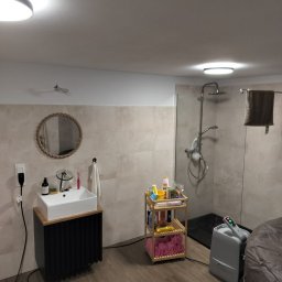 Volodymyr Trufen - Wykończona łazienka z kabiną prysznicową typu walk-in, umywalką nablatową na szafce z lamelami i okrągłym lustrem w ramie ze sznura. Beżowe płytki na ścianach i drewnopodobna podłoga.