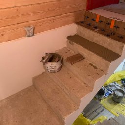 Volodymyr Trufen - Montaż płytek na schodach we Wrocławiu. Widoczny klej, płytki Tubądzin i narzędzia. Częściowo wykończone drewnem ściany i zabezpieczona folią podłoga.