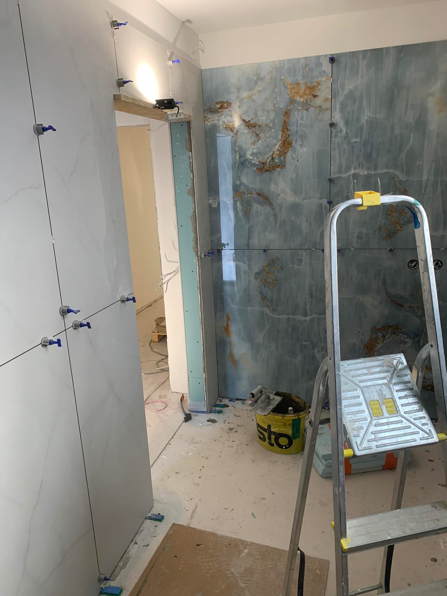 Łazienka w trakcie wykańczania: marmurowe płytki na ścianie z niebieskimi elementami poziomowania, drabina i wiadro z zaprawą. Widoczne narzędzia i surowy stan podłogi.