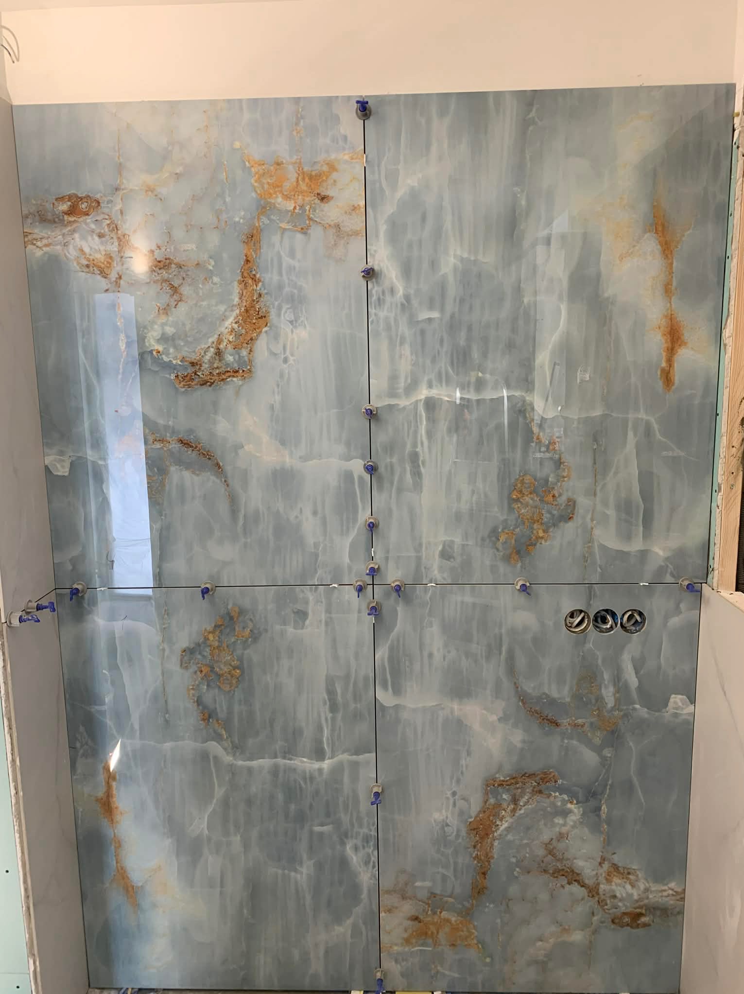 Montaż płytek imitujących marmur w łazience, z widocznymi krzyżykami dystansowymi i otworami na gniazdka elektryczne. Błękitno-złota kolorystyka płytek.