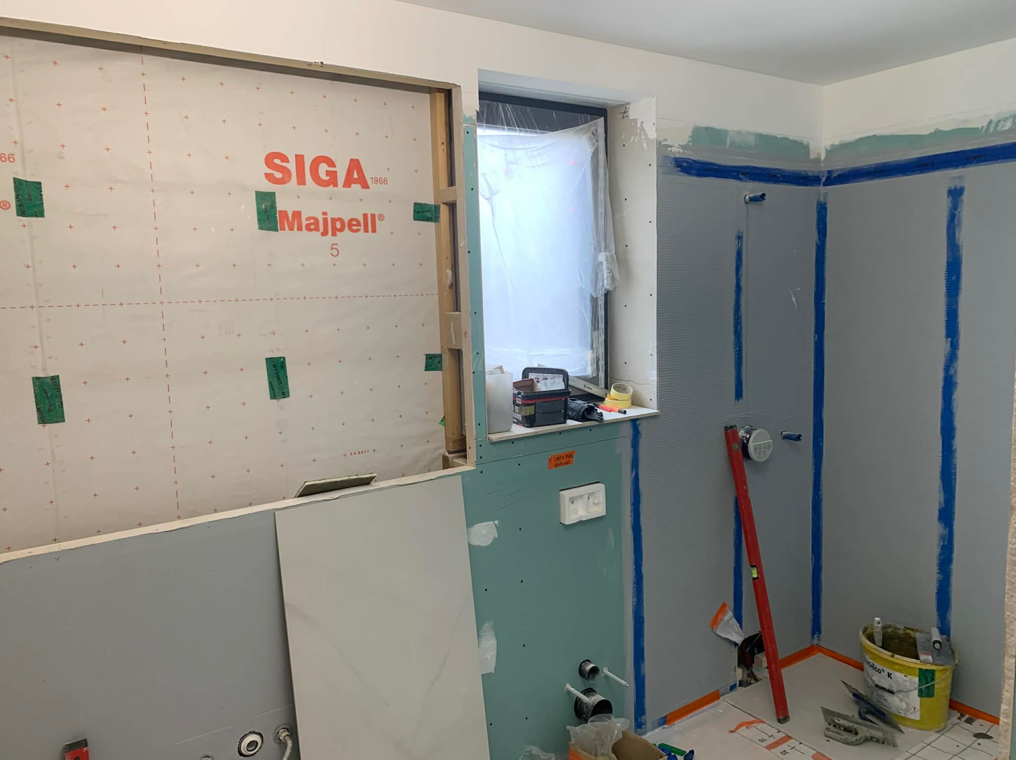 Łazienka w trakcie remontu: ściany z płytami gipsowymi, zabezpieczone folią, okno z folią ochronną, widoczne narzędzia i materiały budowlane, poziomica oparta o ścianę.