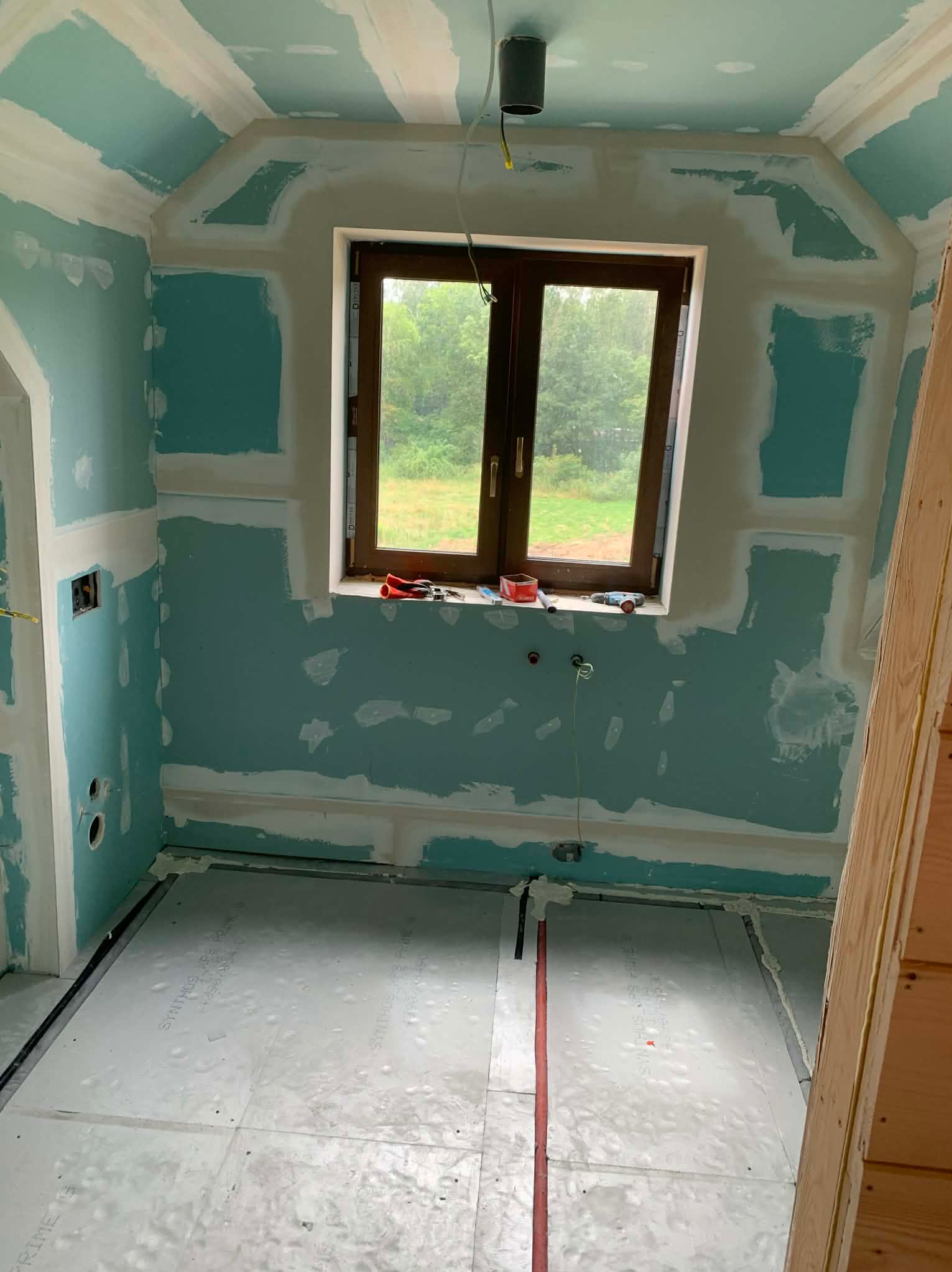 Wnętrze w trakcie prac wykończeniowych: ściany z płyt karton-gips, częściowo wyszpachlowane, okno z narzędziami na parapecie, instalacja podłogowa.