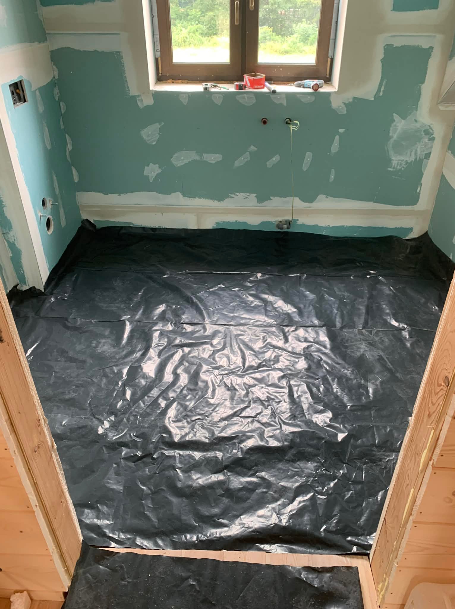 Przygotowanie do remontu: podłoga zabezpieczona czarną folią, ściany z płyt karton-gips, częściowo wyszpachlowane, okno z narzędziami. Wnętrze w trakcie prac.