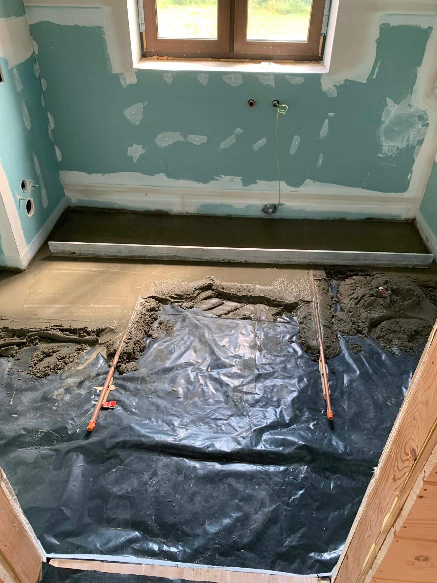 Wyrównywanie wylewki w łazience z niebieskimi ścianami, widoczne narzędzia i folia ochronna. Częściowo wykończone ściany z widocznymi śladami szpachlowania.