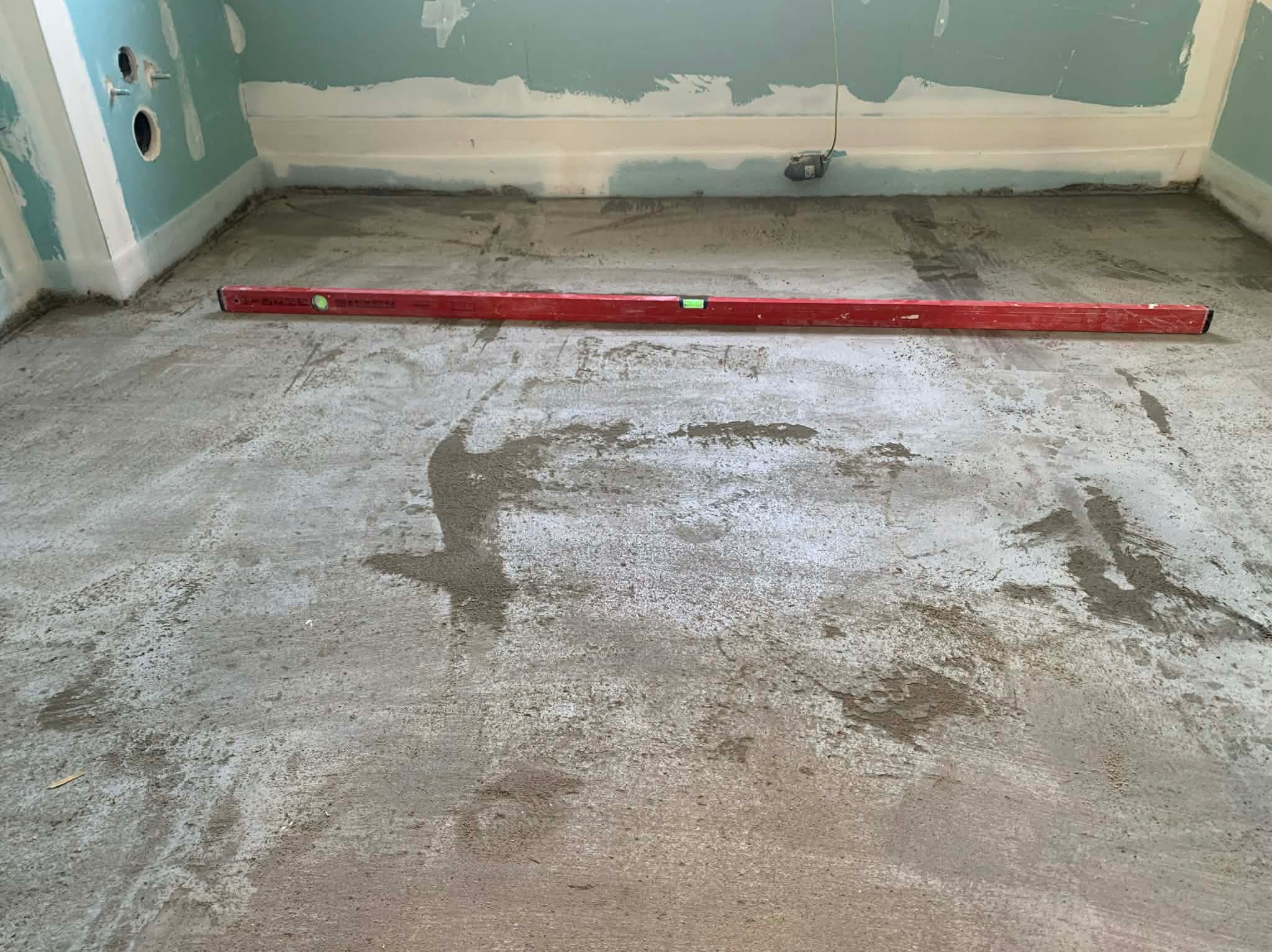 Wyrównywanie podłoża w pomieszczeniu z częściowo wykończonymi ścianami. Czerwona poziomica leży na szarej, mokrej wylewce. Widoczne ślady po narzędziach.