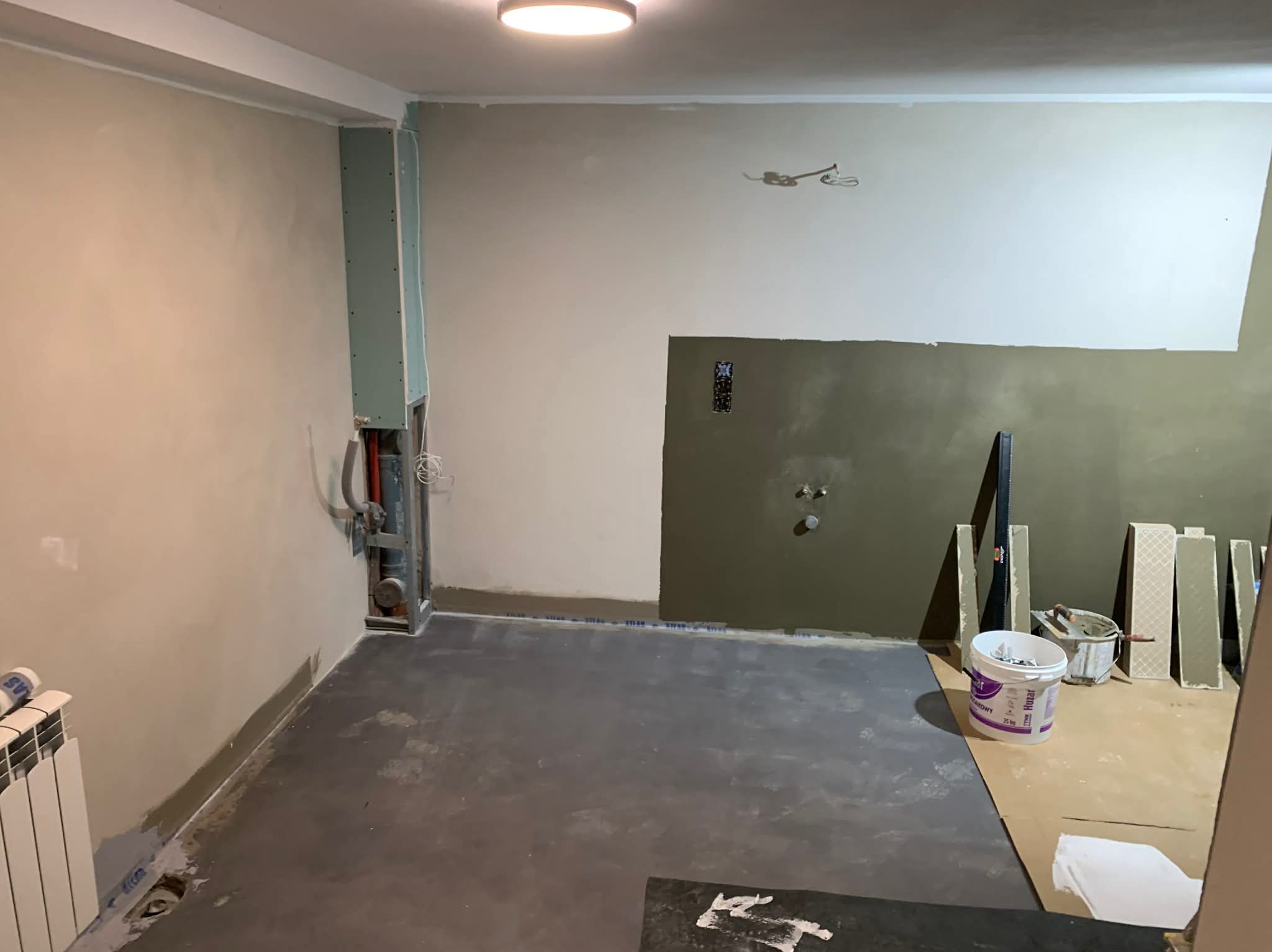 Wnętrze w trakcie remontu: ściany częściowo wyszpachlowane, widoczne instalacje, płyty karton-gips, narzędzia i materiały budowlane. Przygotowanie do dalszych prac wykończeniowych.