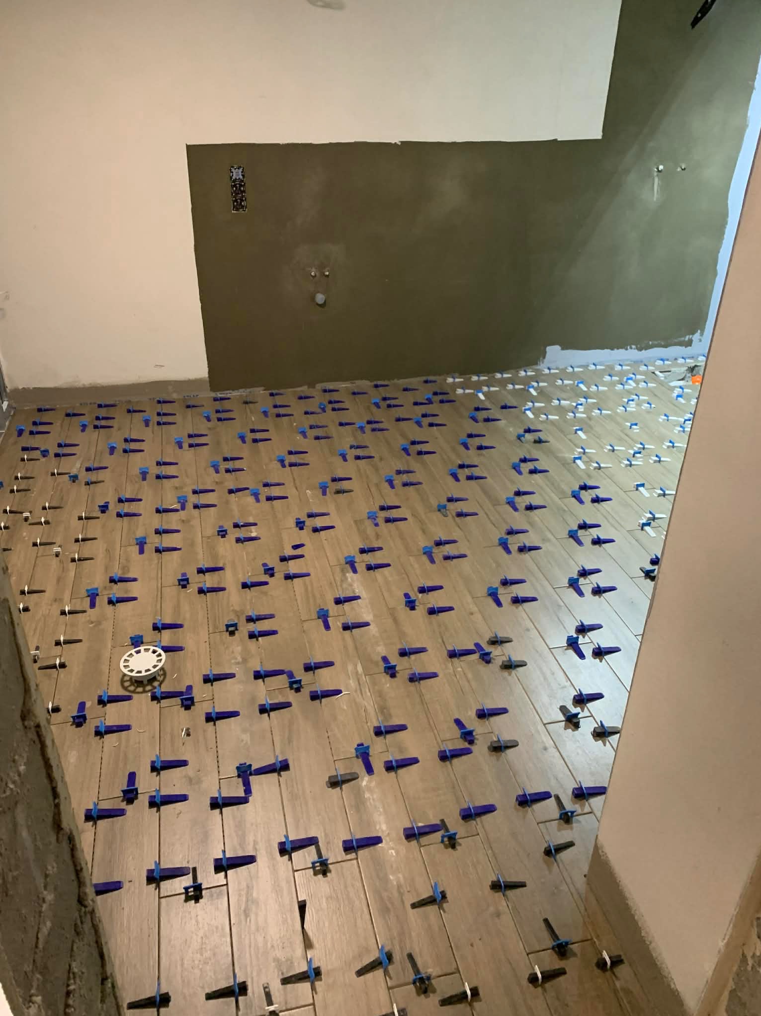 Układanie płytek podłogowych w łazience z użyciem systemu poziomowania. Widoczne krzyżyki dystansowe i kliny. Ściany częściowo przygotowane pod wykończenie.