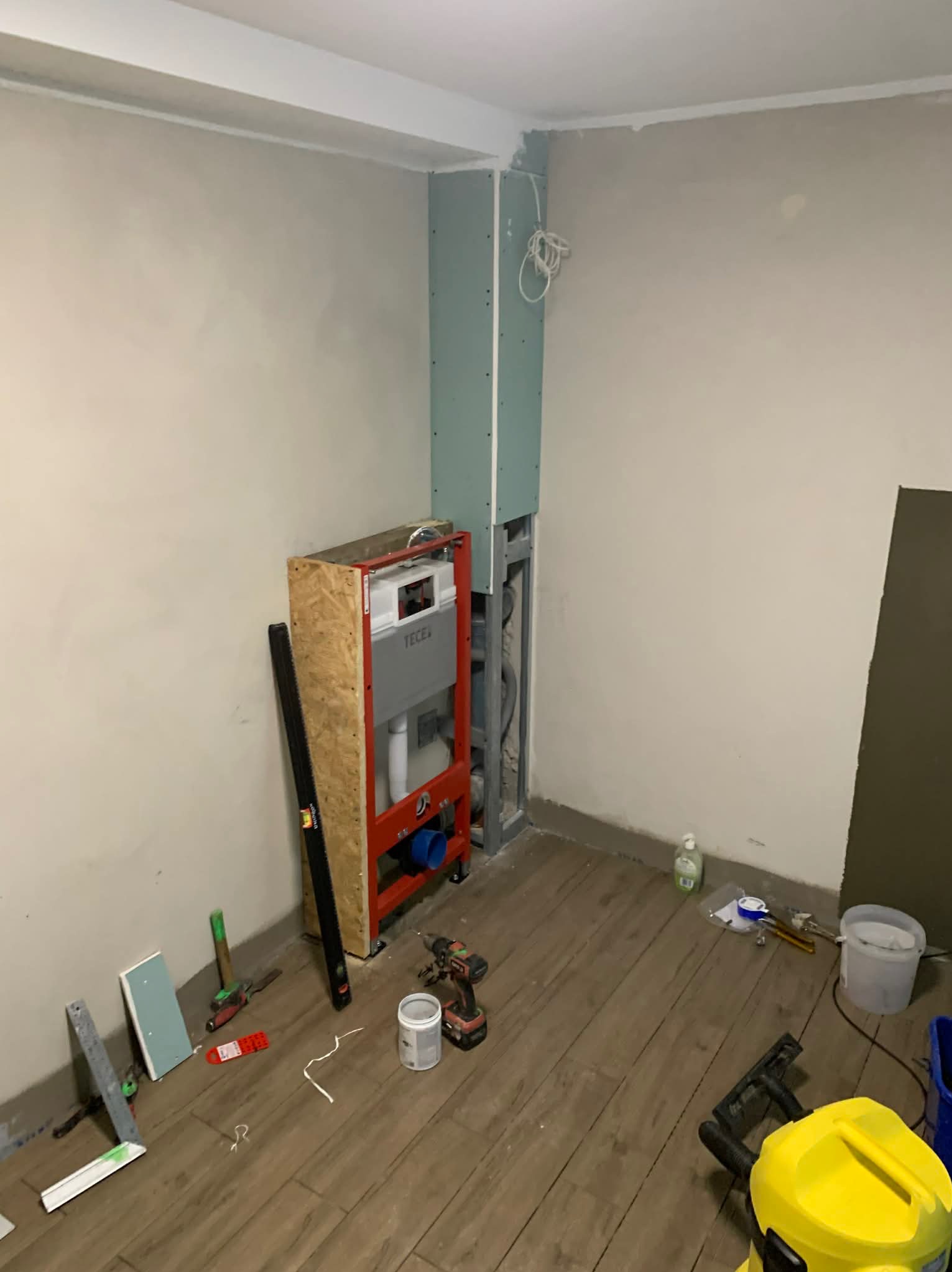 Łazienka w trakcie remontu: stelaż podtynkowy WC, płyta karton-gips, narzędzia na podłodze imitującej drewno. Widoczne elementy instalacji.