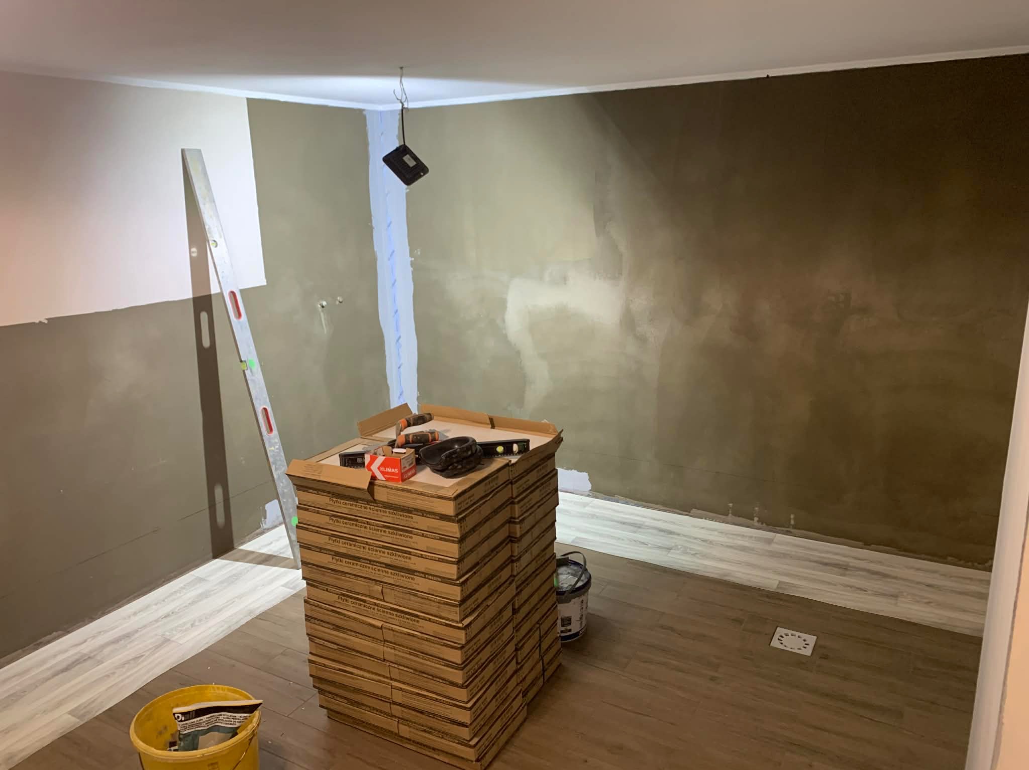 Wnętrze w trakcie wykańczania: ułożone płytki podłogowe imitujące drewno, sterta kartonów z płytkami ściennymi, poziomica i narzędzia. Ściany częściowo wyszpachlowane.