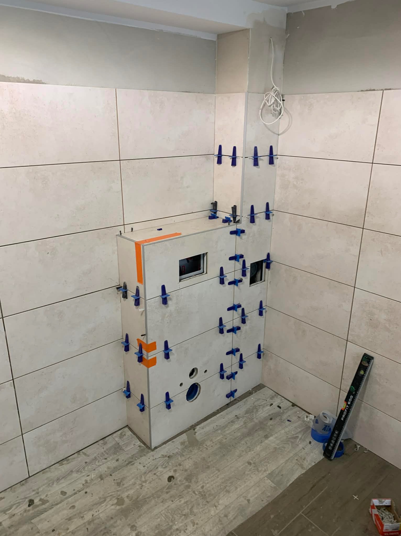 Montaż płytek w łazience, widoczna zabudowa stelaża WC z użyciem klipsów poziomujących, poziomica i resztki po pracy na podłodze. Jasne płytki na ścianach.