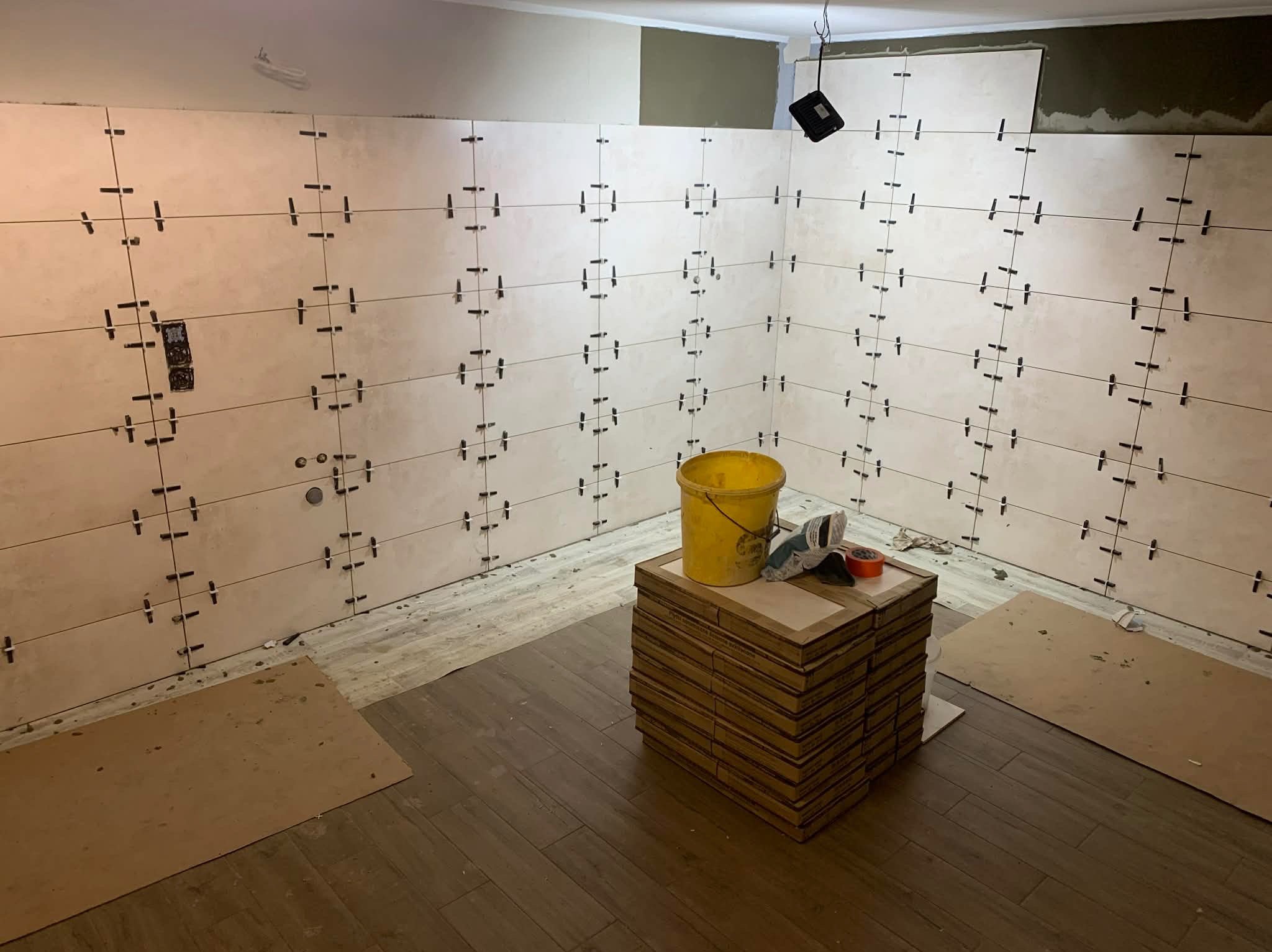 Ściany w trakcie układania płytek z użyciem krzyżyków dystansowych, widoczny żółty kubeł i kartony z płytkami na podłodze imitującej drewno.