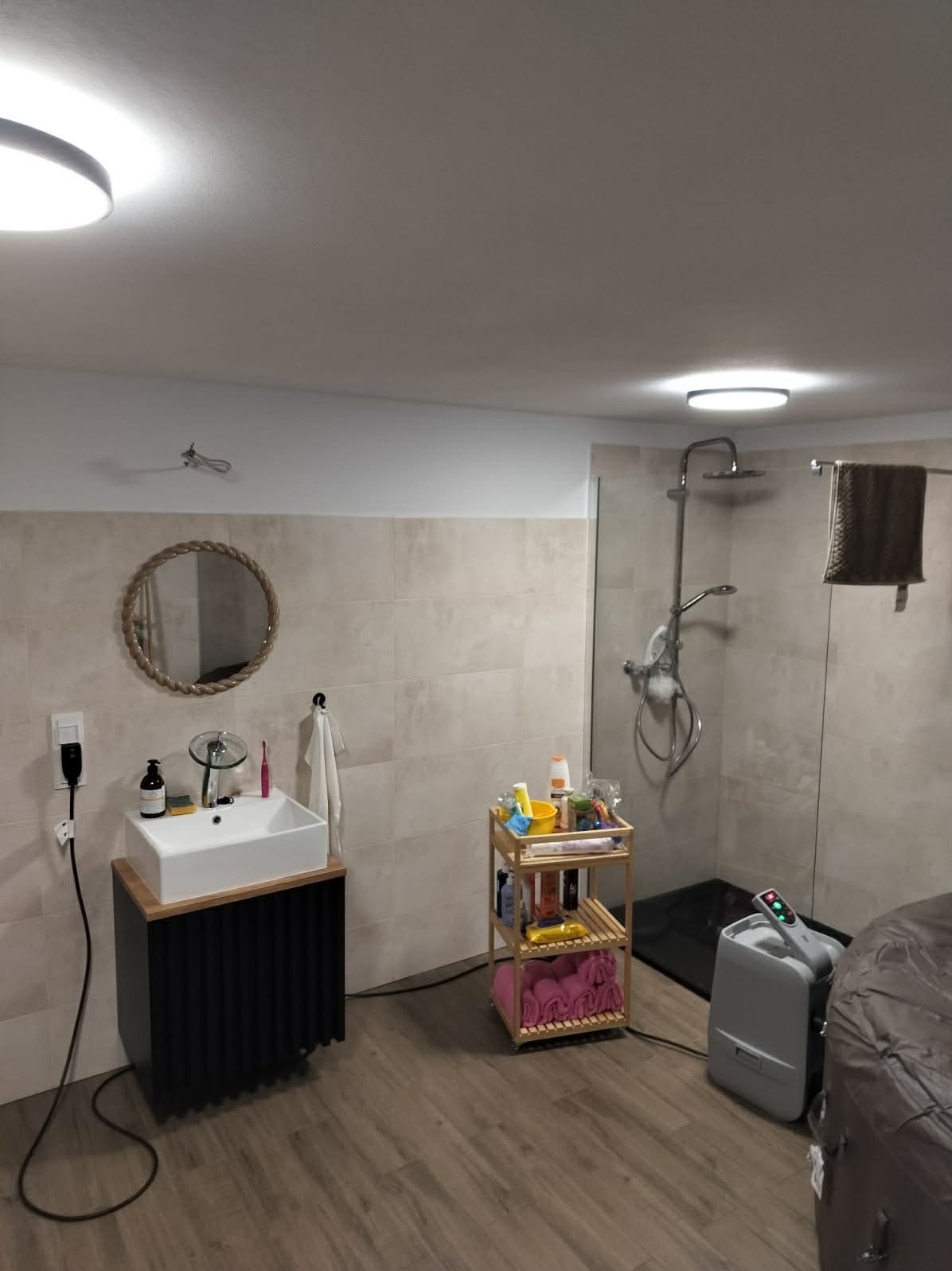 Wykończona łazienka z kabiną prysznicową typu walk-in, umywalką nablatową na szafce z lamelami i okrągłym lustrem w ramie ze sznura. Beżowe płytki na ścianach i drewnopodobna podłoga.