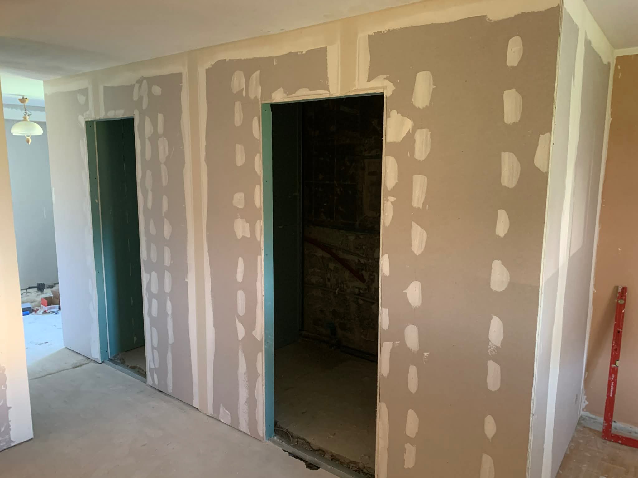 Montaż płyt karton-gips na ścianach z widocznymi łączeniami i szpachlowaniem. W tle widoczne otwory drzwiowe i poziomica oparta o ścianę.