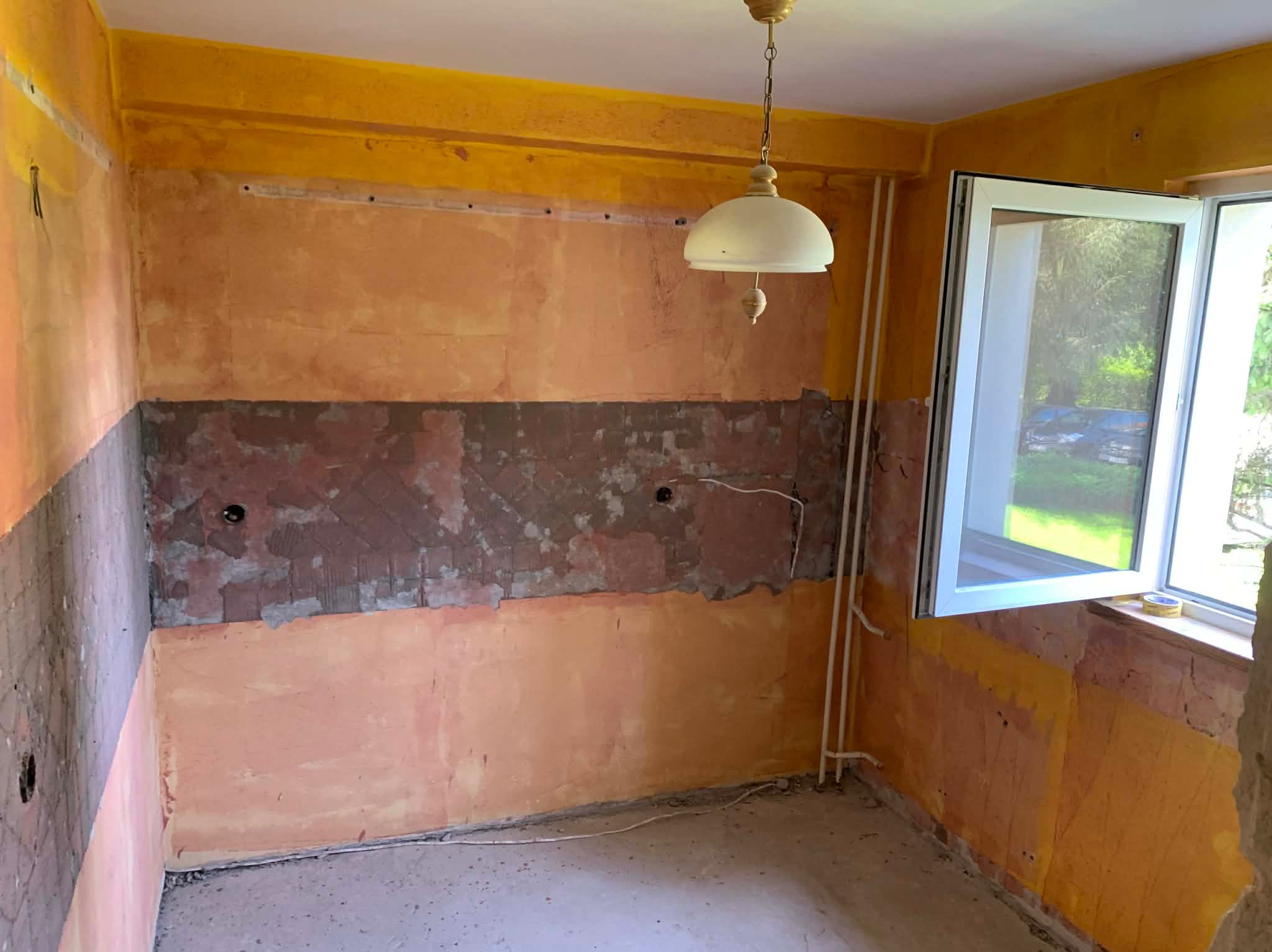 Wnętrze kuchni w trakcie remontu: ściany z fragmentami starej farby, odsłonięty fragment muru, nowe okno PCV, wisząca lampa. Widoczne ślady po instalacjach.