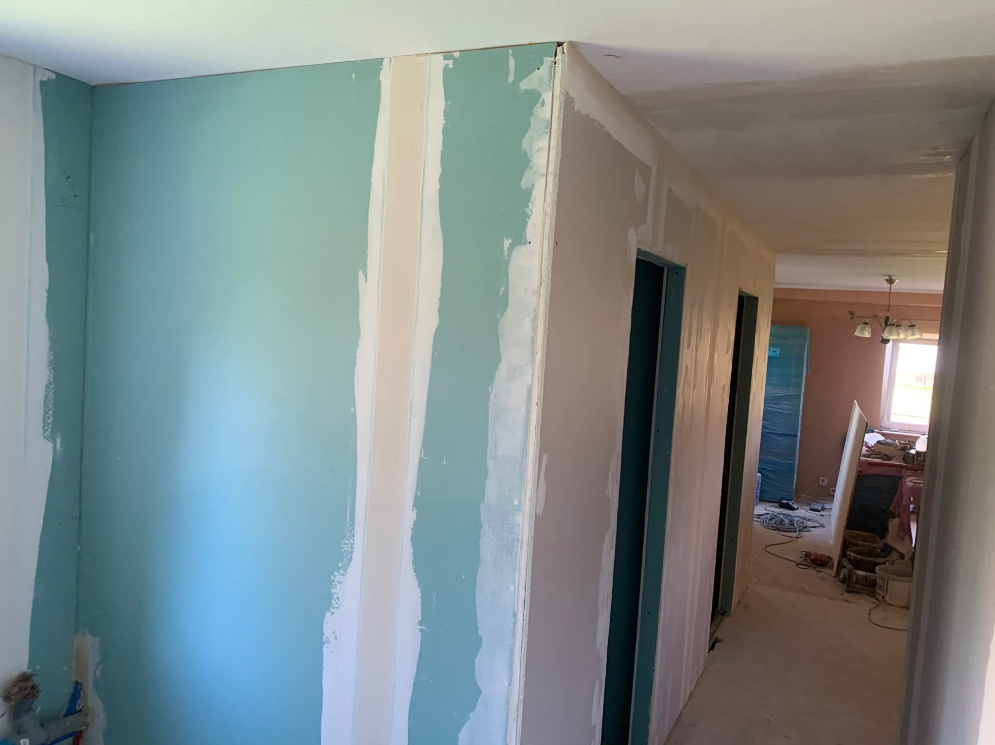Ściana z płyt karton-gips, częściowo pomalowana na turkusowo, z widocznymi łączeniami i szpachlowaniem. W tle widoczne dalsze pomieszczenia w trakcie remontu.