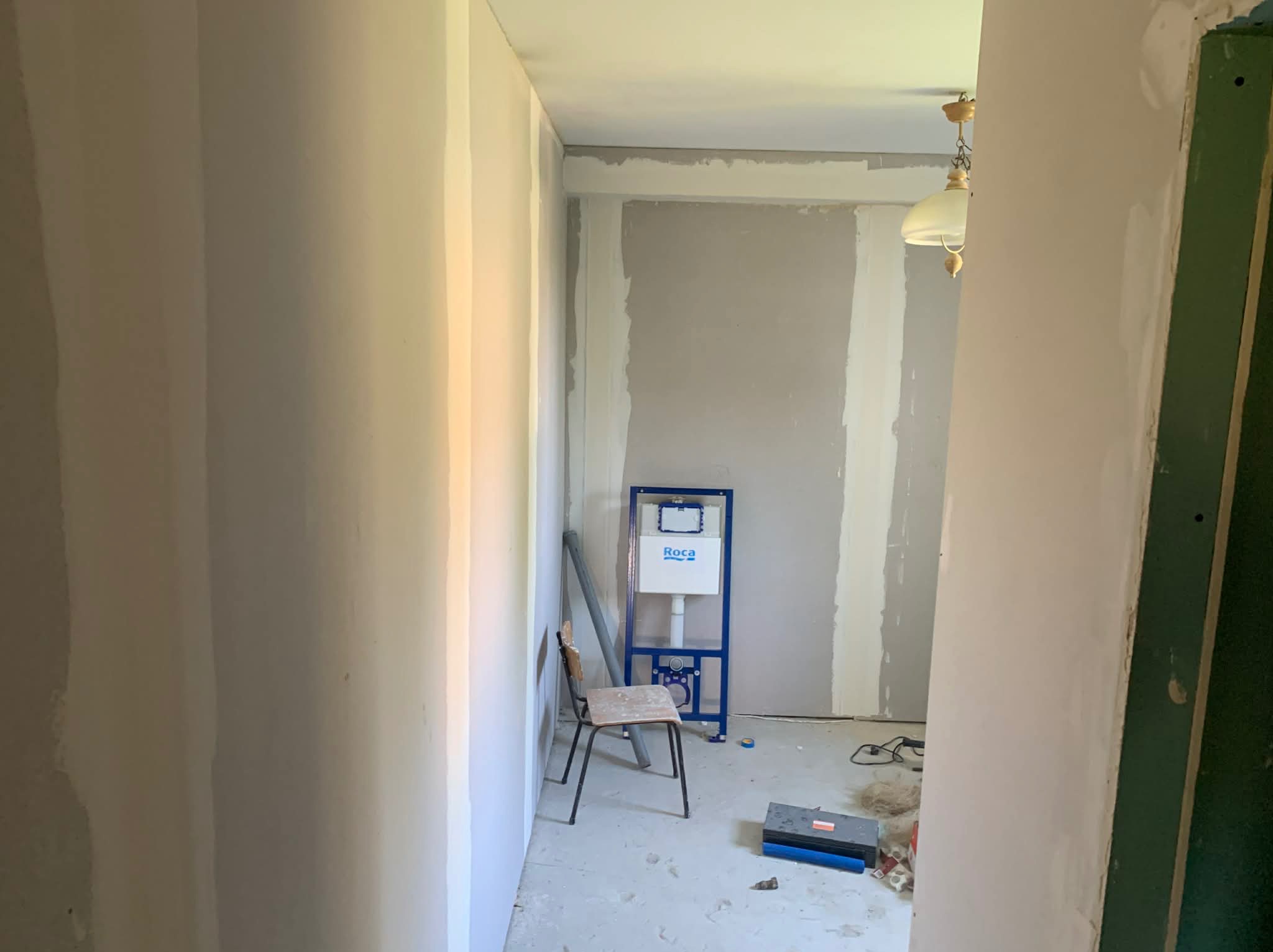 Wnętrze w trakcie remontu: ściany po szpachlowaniu, stelaż podtynkowy WC Roca, krzesło i narzędzia na podłodze. Widok z korytarza, surowy stan wykończenia.
