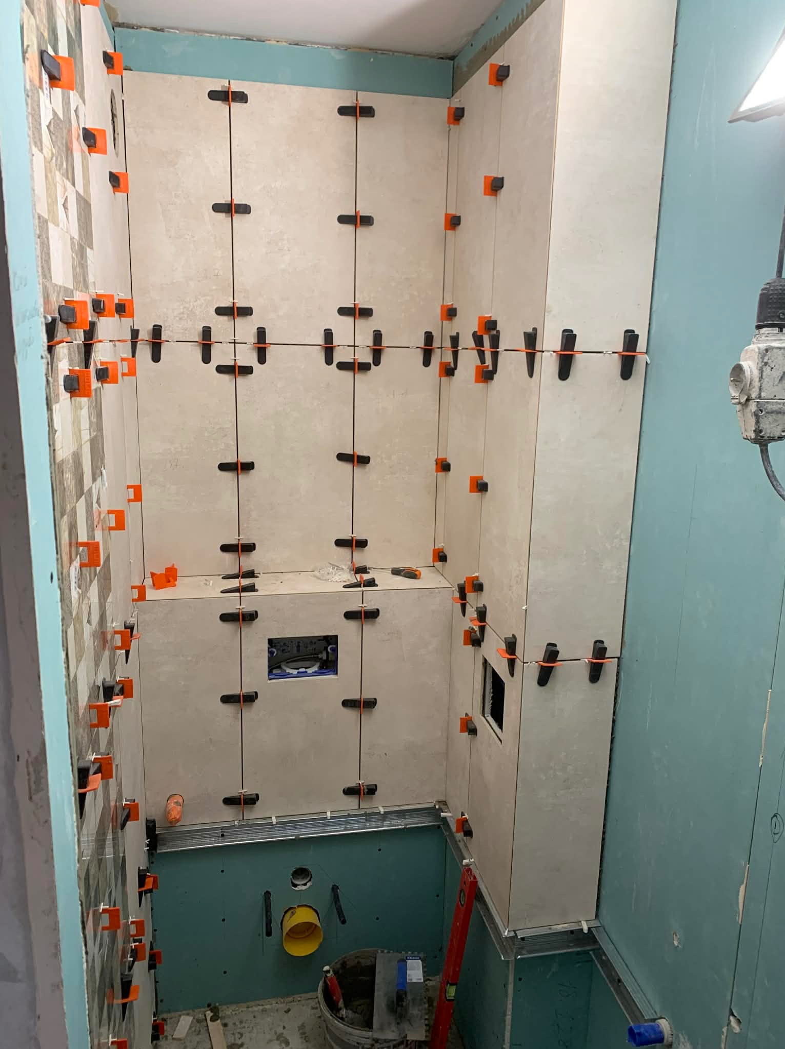 Wnętrze łazienki w trakcie układania płytek. Widoczne płytki w odcieniach beżu i szarości, krzyżyki dystansowe i poziomica. Częściowo wykończona ściana z mozaiką.