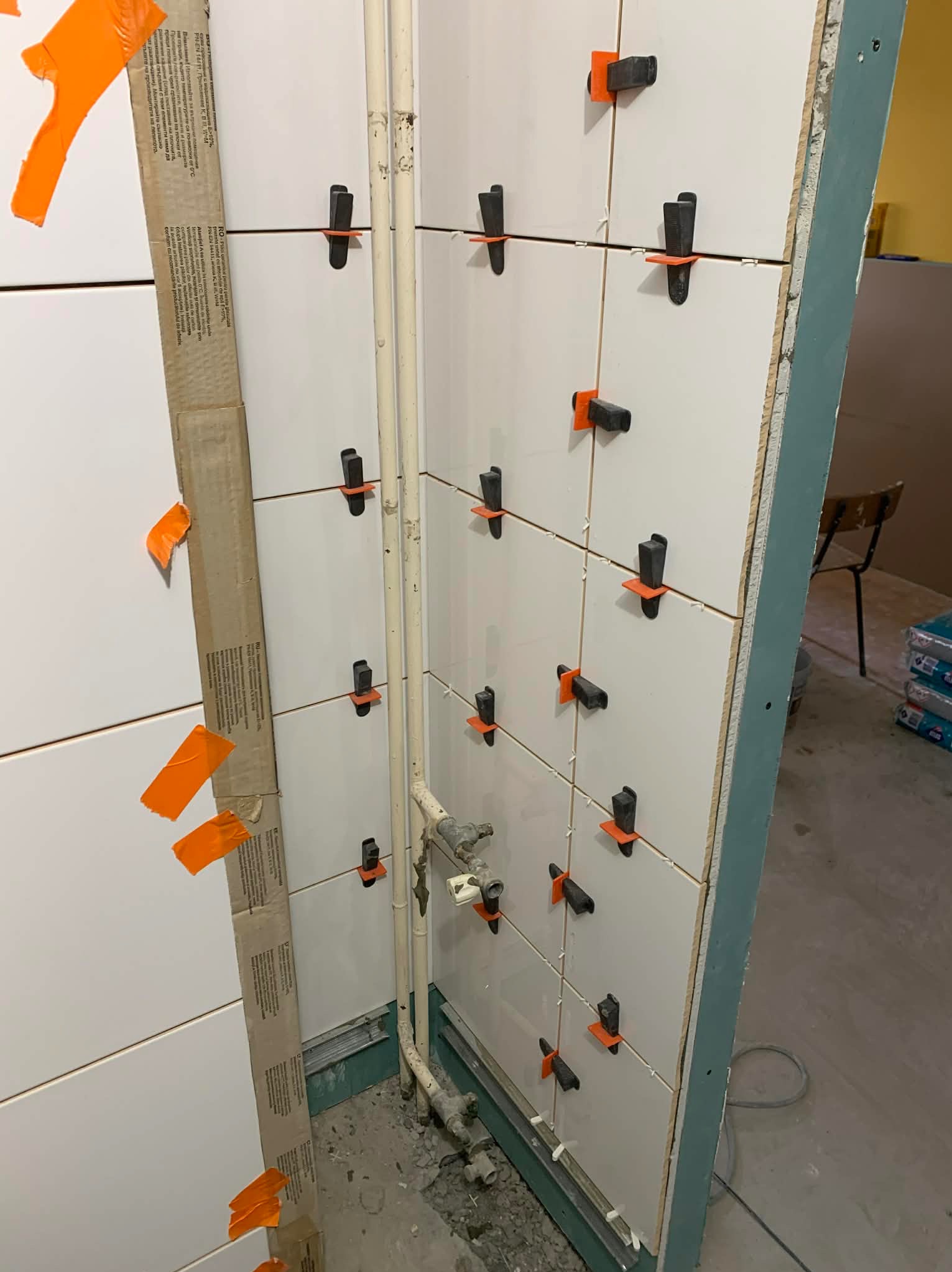 Białe płytki ścienne w trakcie montażu, z użyciem klipsów poziomujących, widoczne rury instalacyjne i karton z instrukcją. Detal z wykończenia łazienki.