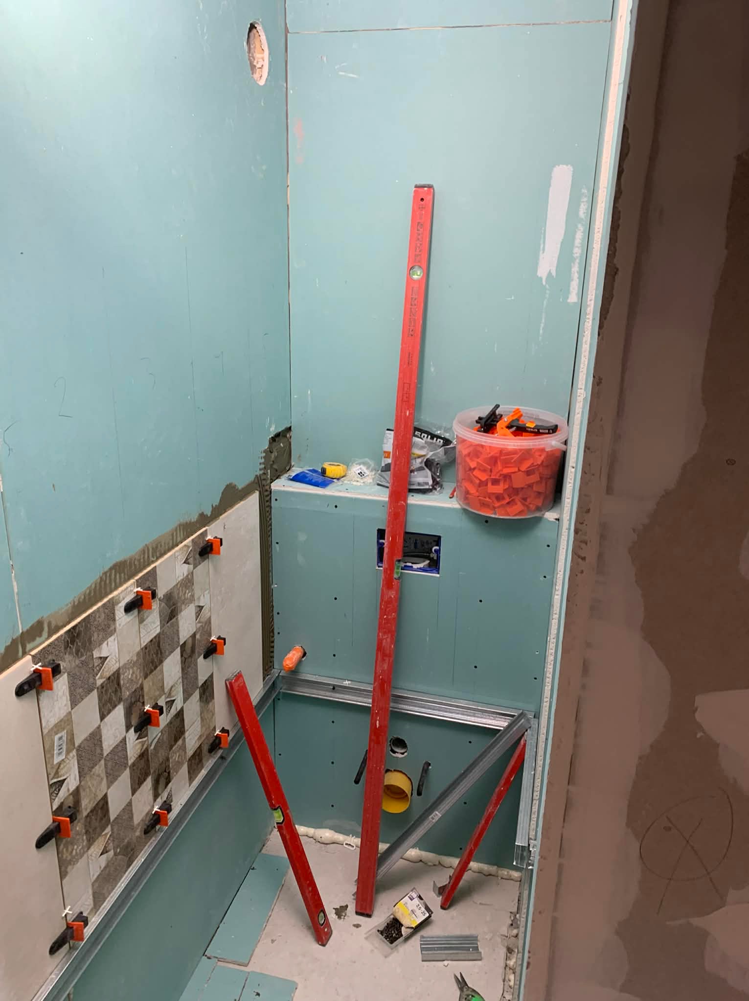 Łazienka w trakcie wykańczania: częściowo ułożone płytki w szachownicę, poziomice, płyta karton gips, narzędzia i akcesoria do montażu płytek.