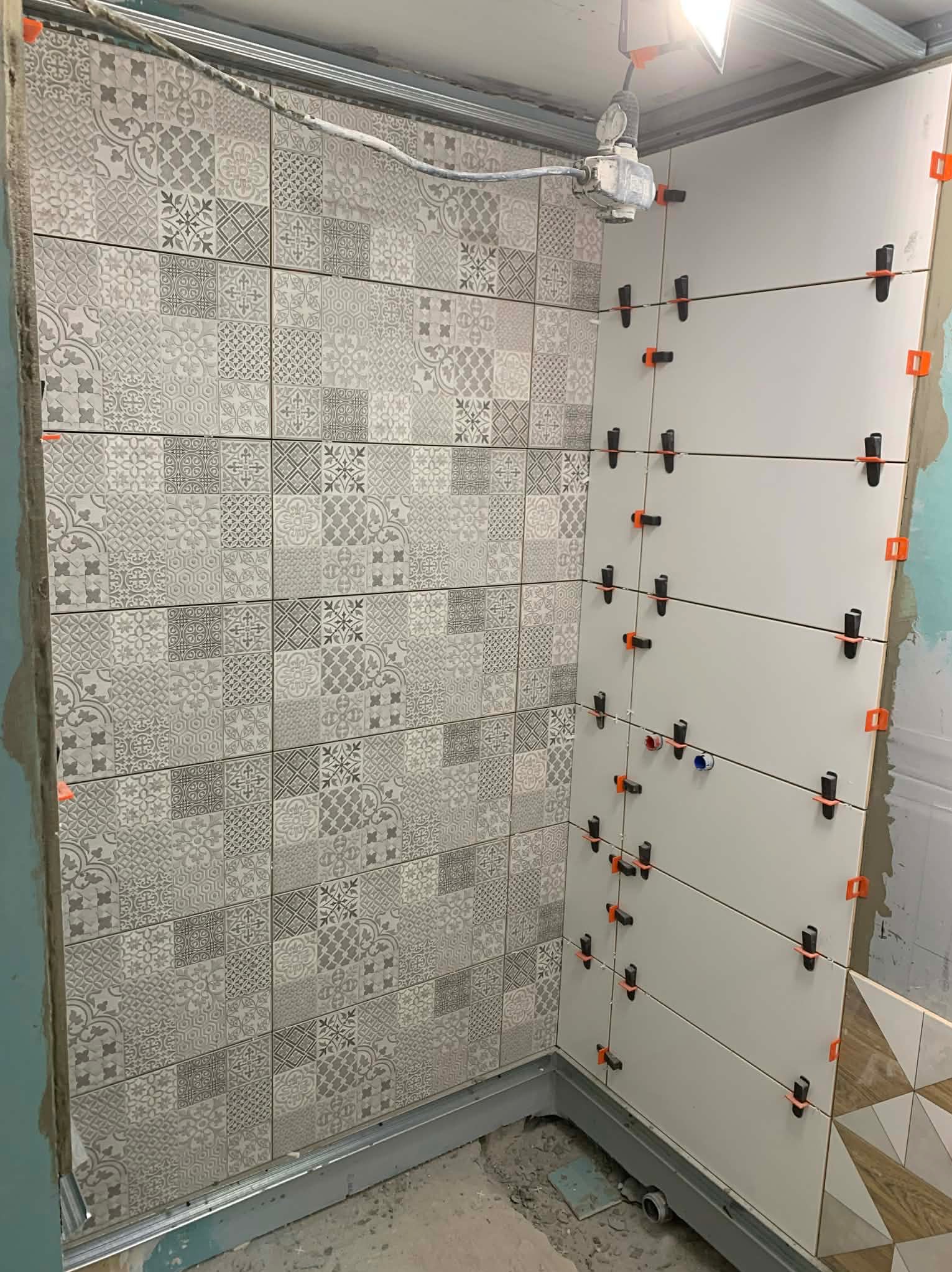 Układanie płytek w łazience z użyciem klipsów poziomujących. Ściana wyłożona wzorzystymi, szarymi płytkami, druga ściana z jasnymi, gładkimi płytkami. Widoczna lampa robocza.