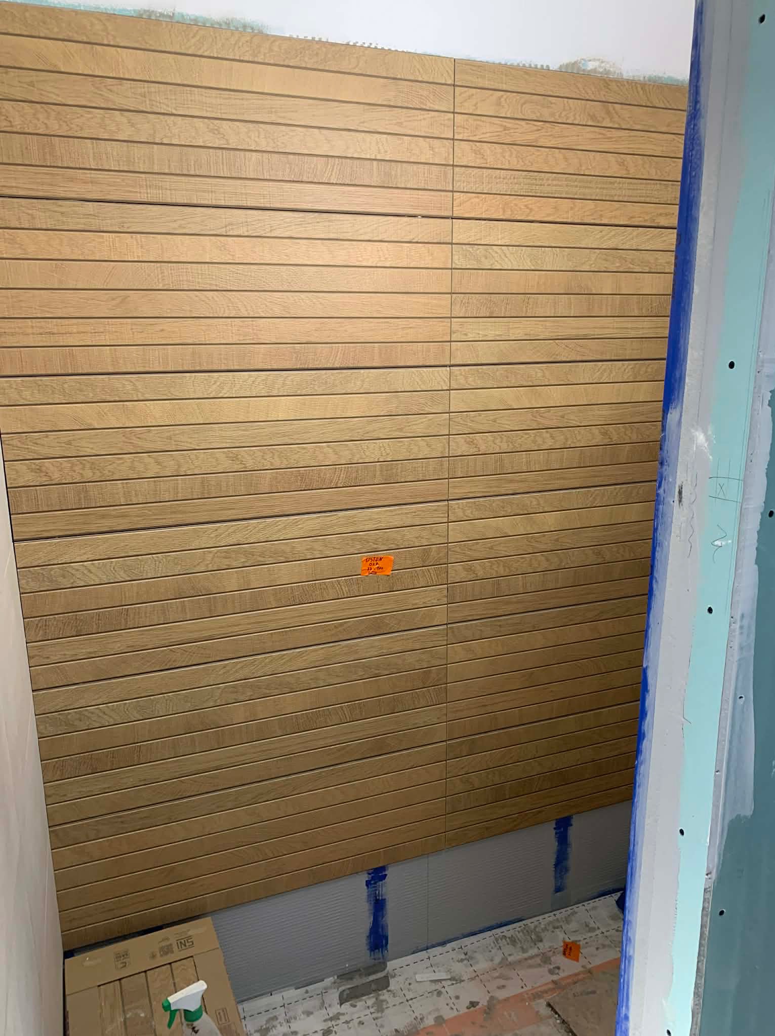 Ściana wyłożona podłużnymi płytkami imitującymi drewno w łazience, z widocznymi elementami montażowymi i kartonem na podłodze. Płytki ułożone poziomo.