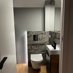 EkoPainter - Nowoczesna łazienka z marmurową ścianą, podświetleniem LED, wiszącą toaletą i umywalką z szafką w odcieniach drewna. Widok zza uchylonych drzwi.
