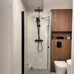 EkoPainter - Nowoczesna łazienka z kabiną typu walk-in, czarną armaturą i drewnianą zabudową. Minimalistyczny design, jasne płytki i oświetlenie sufitowe.