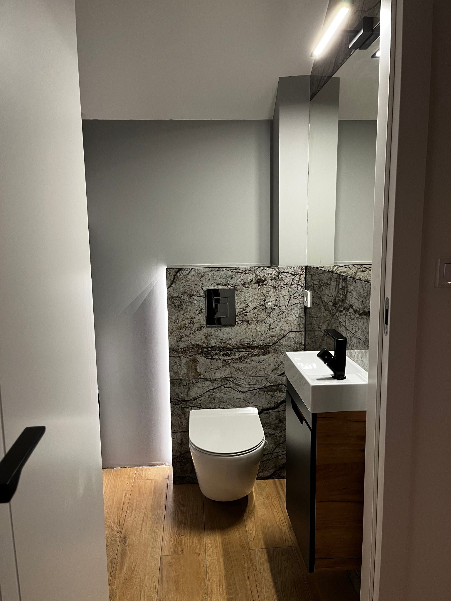 Nowoczesna łazienka z marmurową ścianą, podświetleniem LED, wiszącą toaletą i umywalką z szafką w odcieniach drewna. Widok zza uchylonych drzwi.