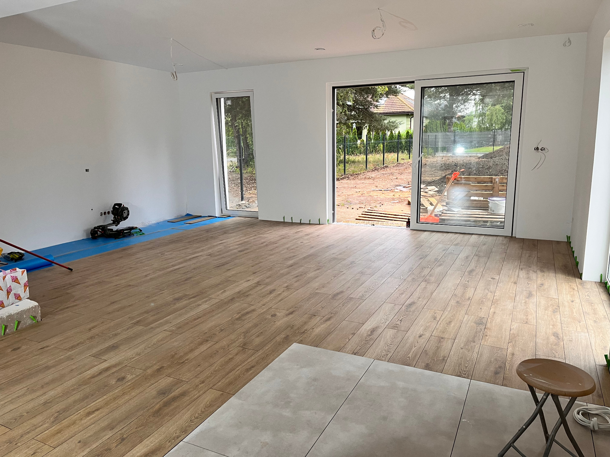 Jasne panele podłogowe imitujące drewno w przestronnym wnętrzu z białymi ścianami i dużymi oknami. Widoczne narzędzia i materiały budowlane.