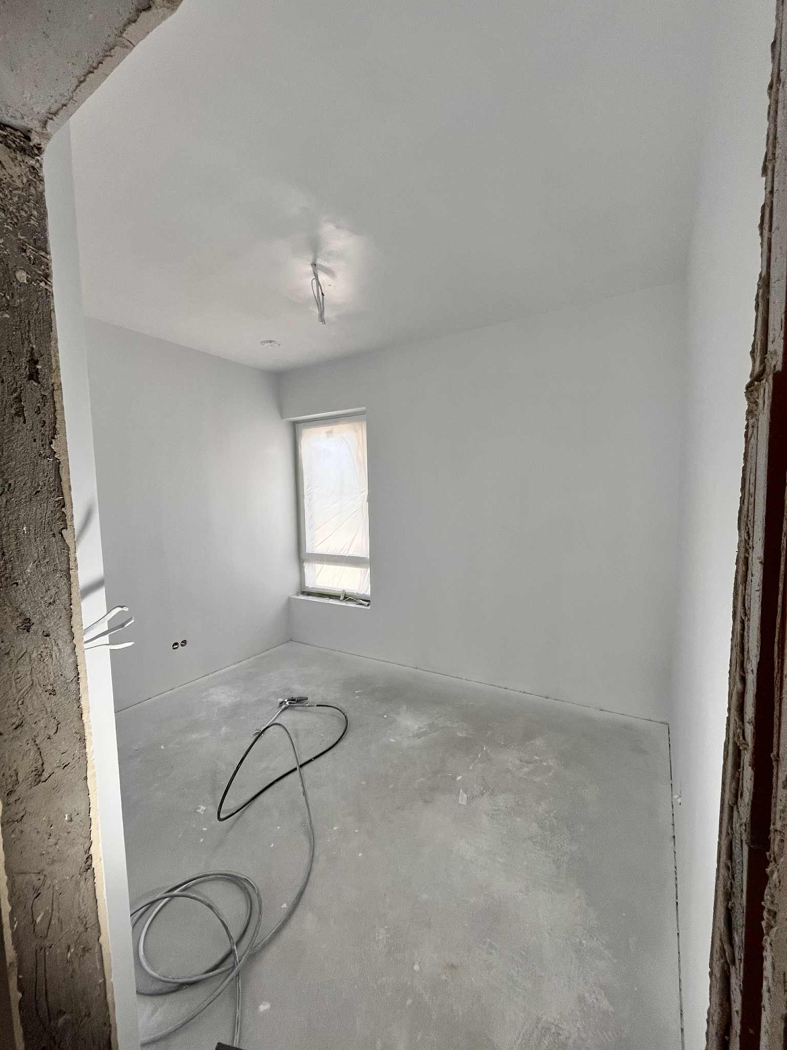 Wnętrze pokoju po szpachlowaniu i malowaniu na biało, z widocznym sprzętem malarskim na podłodze i zabezpieczonym oknem folią malarską. Surowy stan wykończenia ościeżnic.