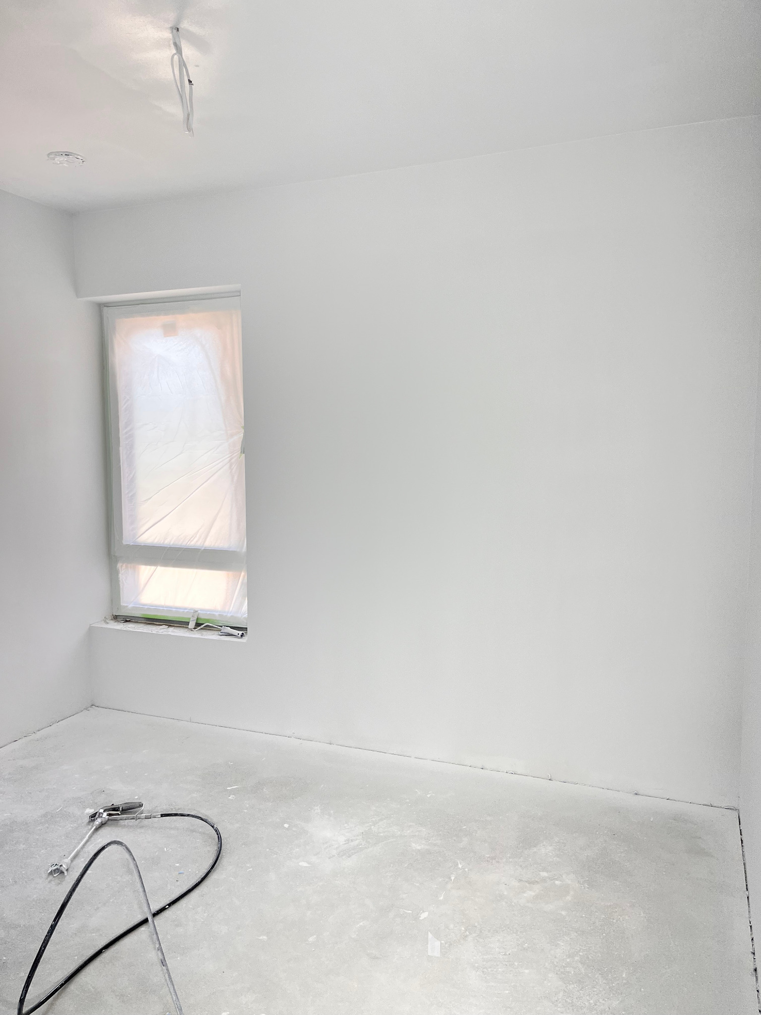 Białe, świeżo pomalowane wnętrze z zakrytym oknem i sprzętem do malowania natryskowego na podłodze. Minimalistyczny kadr, jasne oświetlenie, surowy beton na podłodze.