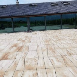 Greks-Bud - Nowoczesny taras z geometryczną kostką w odcieniach beżu i brązu, duże okna domu z odbiciem krajobrazu w tle, błękitne niebo. Widok z perspektywy.