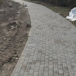 Greks-Bud - Ścieżka z szarej kostki brukowej wijąca się przez las, z widocznymi śladami prac ziemnych po bokach i workiem w tle. Ujęcie z perspektywy pieszego.