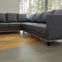 X-Mara Dzianis Khmara - Szary, narożny, tapicerowany zestaw wypoczynkowy z poduszkami, na drewnianej ramie, z niskim oparciem, na tle okna z widokiem na las i parkiet w jodełkę.