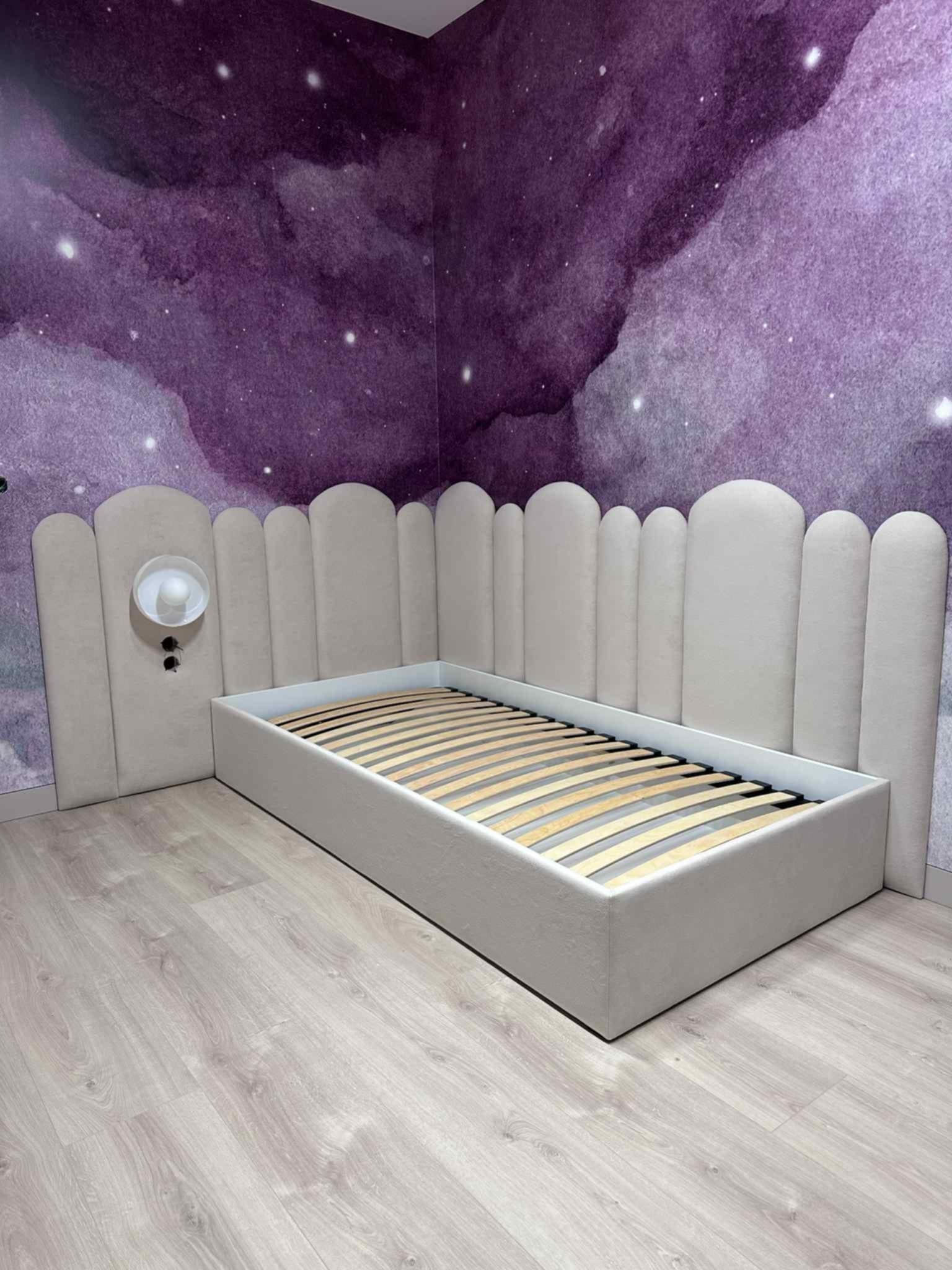 Tapicerowane łóżko z wezgłowiem w kształcie zaokrąglonych paneli, z lampką nocną, na tle ściany z motywem kosmosu. Jasna podłoga z paneli.