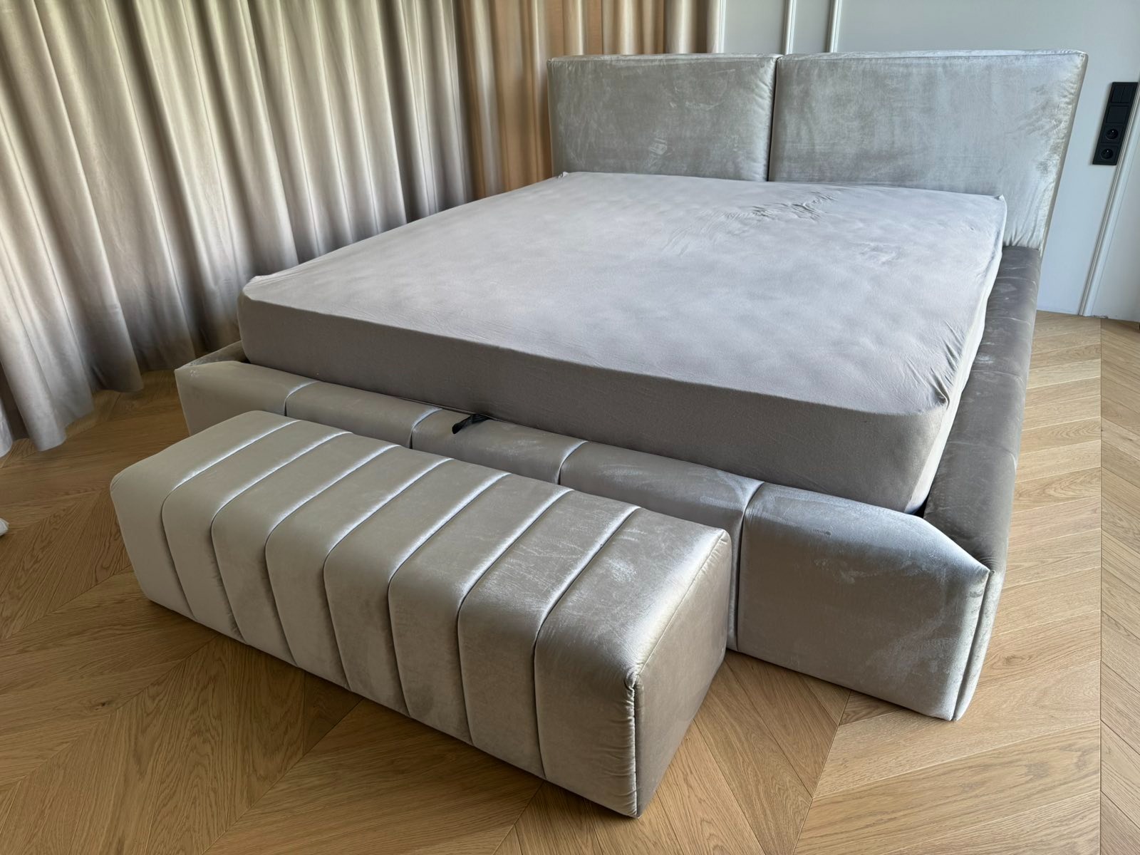 Tapicerowane łóżko z wezgłowiem i pufą w odcieniach szarości, na parkiecie ułożonym w jodełkę. Minimalistyczny design, miękkie światło, zasłony w tle.