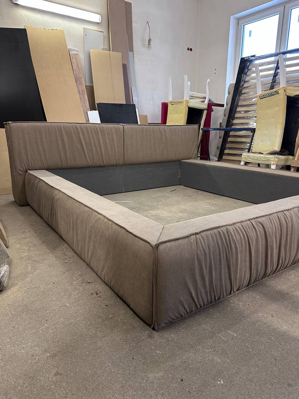 Tapicerowane łóżko w odcieniach beżu, widoczne w warsztacie. Rama z charakterystycznym marszczeniem materiału. W tle płyty meblowe i elementy stelaża.