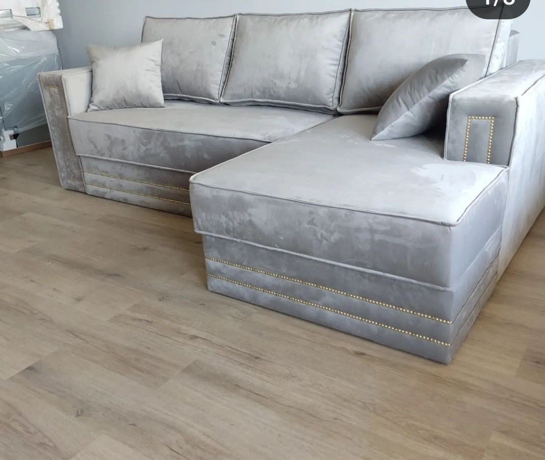 Szary, welurowy narożnik z ozdobnymi nitami, ustawiony na drewnianej podłodze. Nowoczesny design, miękkie poduszki, eleganckie wykończenie.